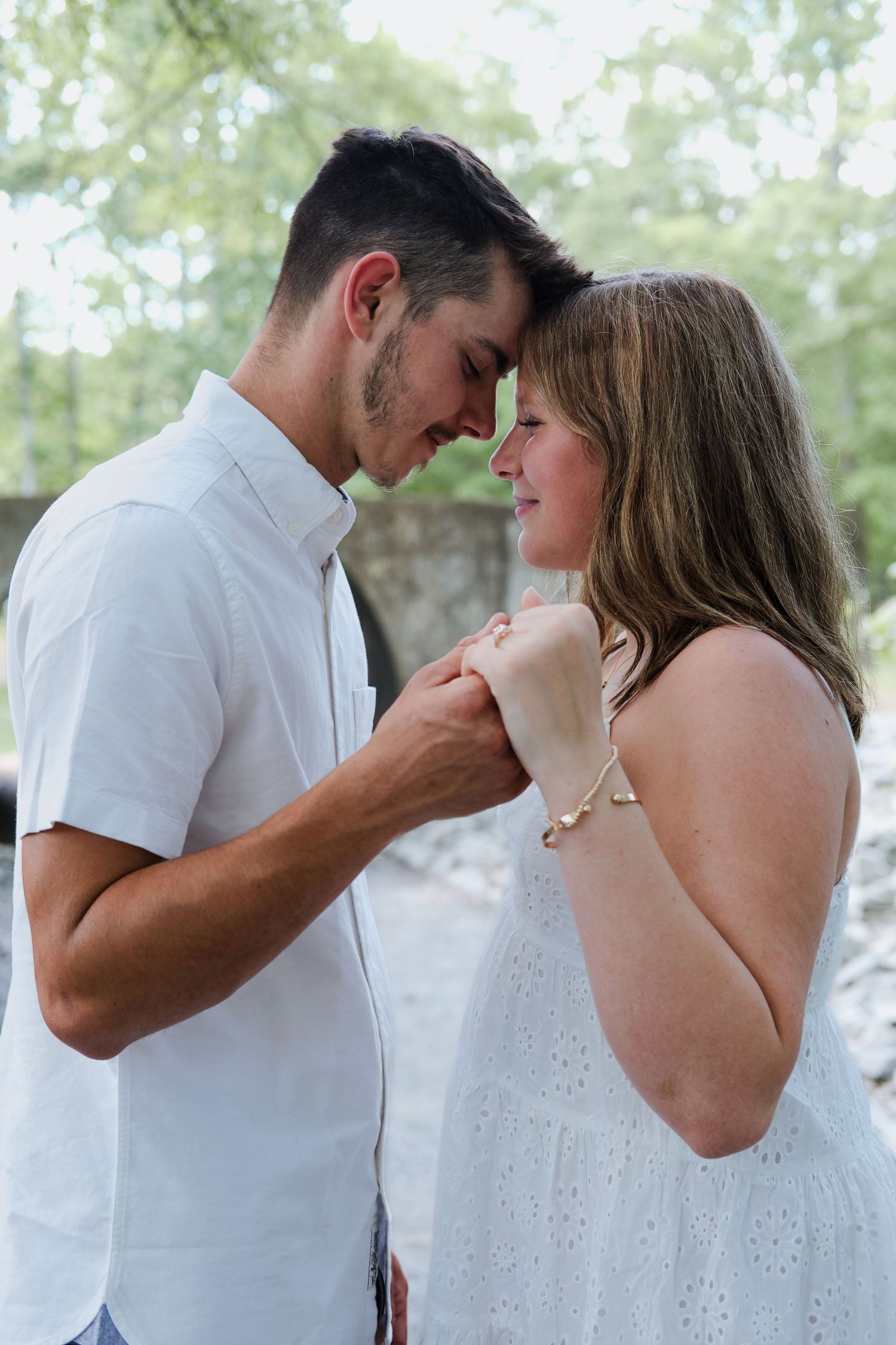 A romantic couple in white outfits touching foreheads and holding hands outdoors with lush green trees in the background.