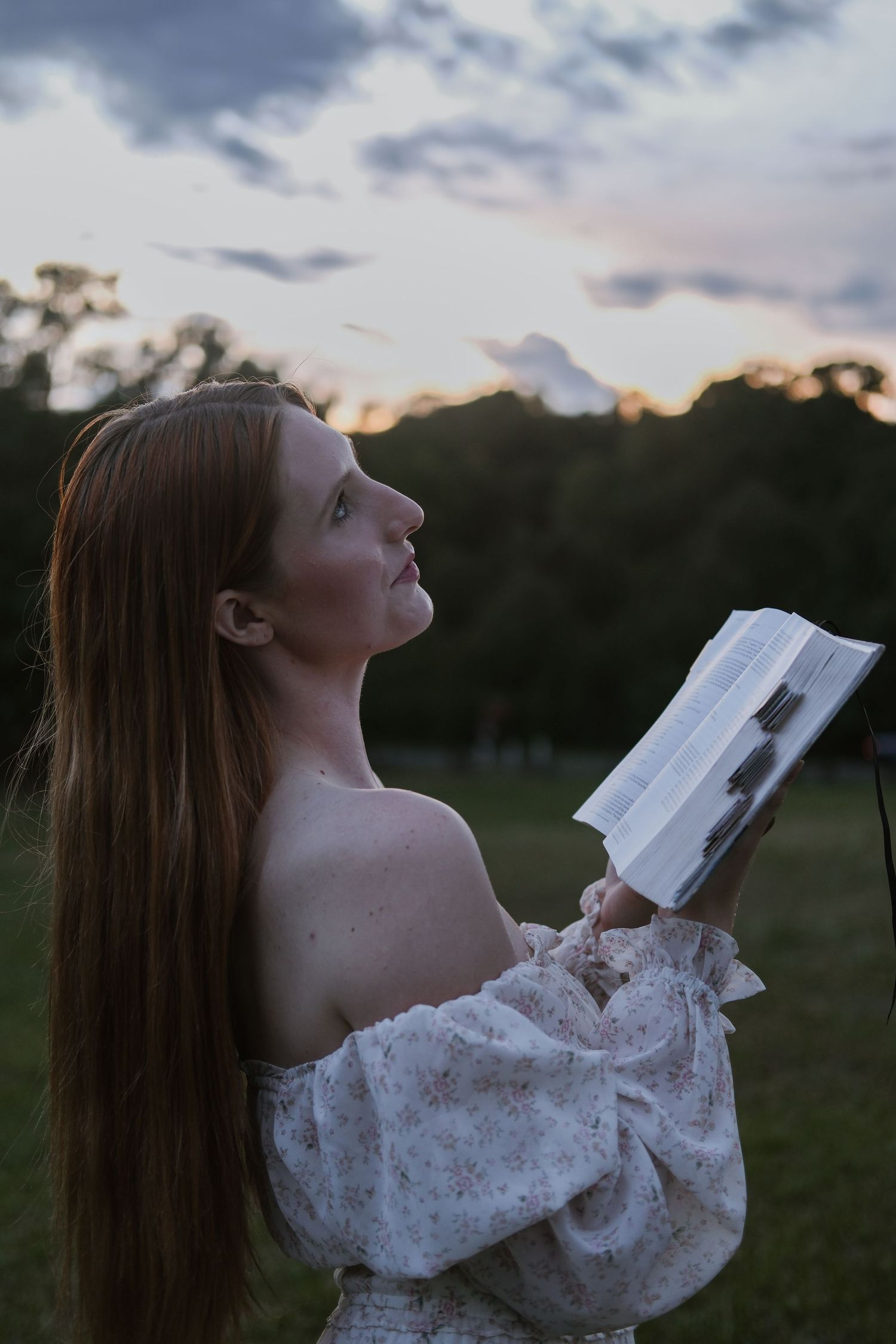 A young redhead woman in a floral off-shoulder dress holds a book while gazing upward at a dramatic sunset sky.