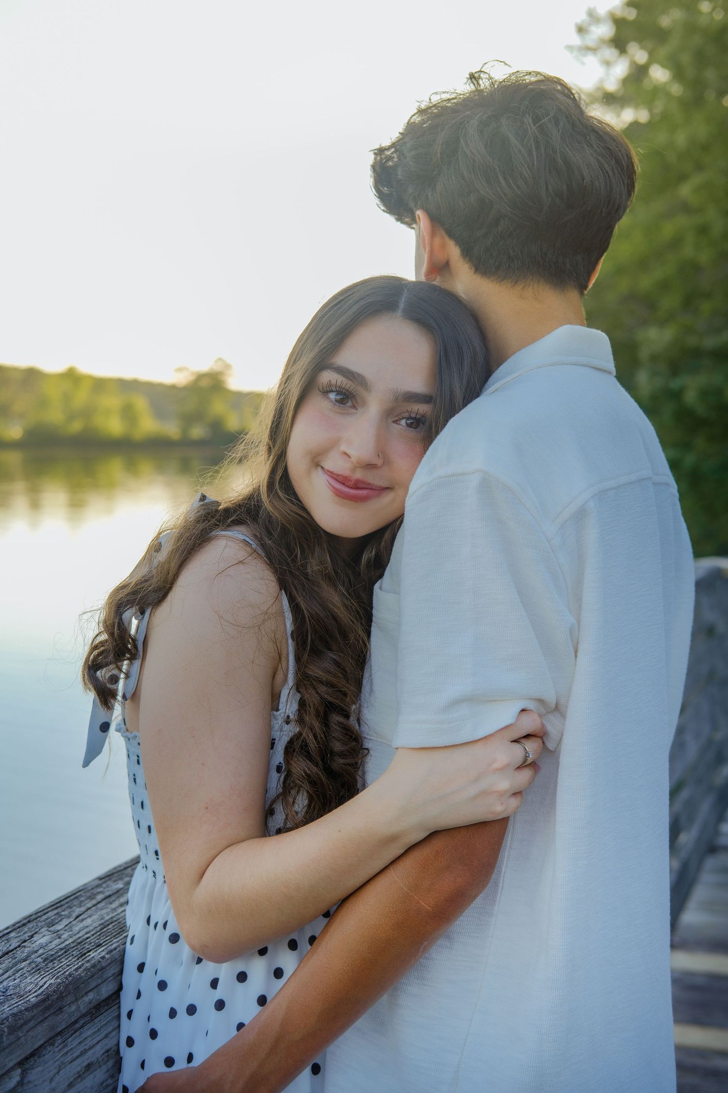 Young woman smiles at camera while hugging her boyfriend on a lakeside dock at dusk.