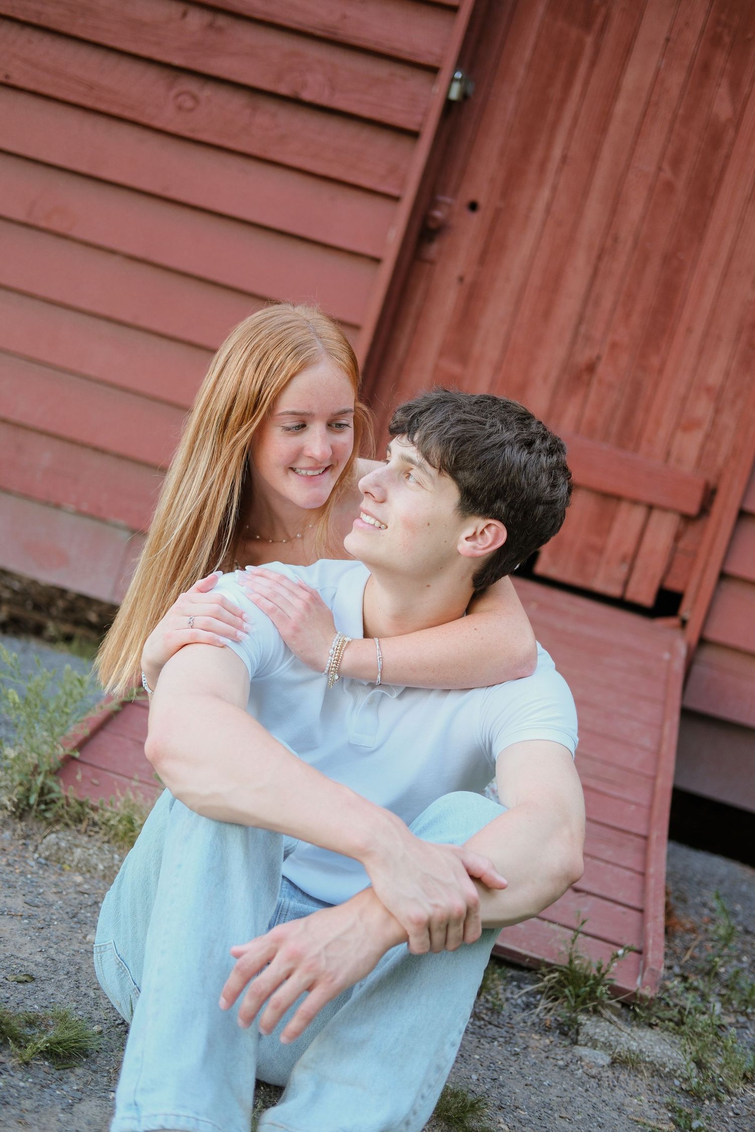 A young couple sitting together outdoors, smiling and embracing near a red wooden barn wall.