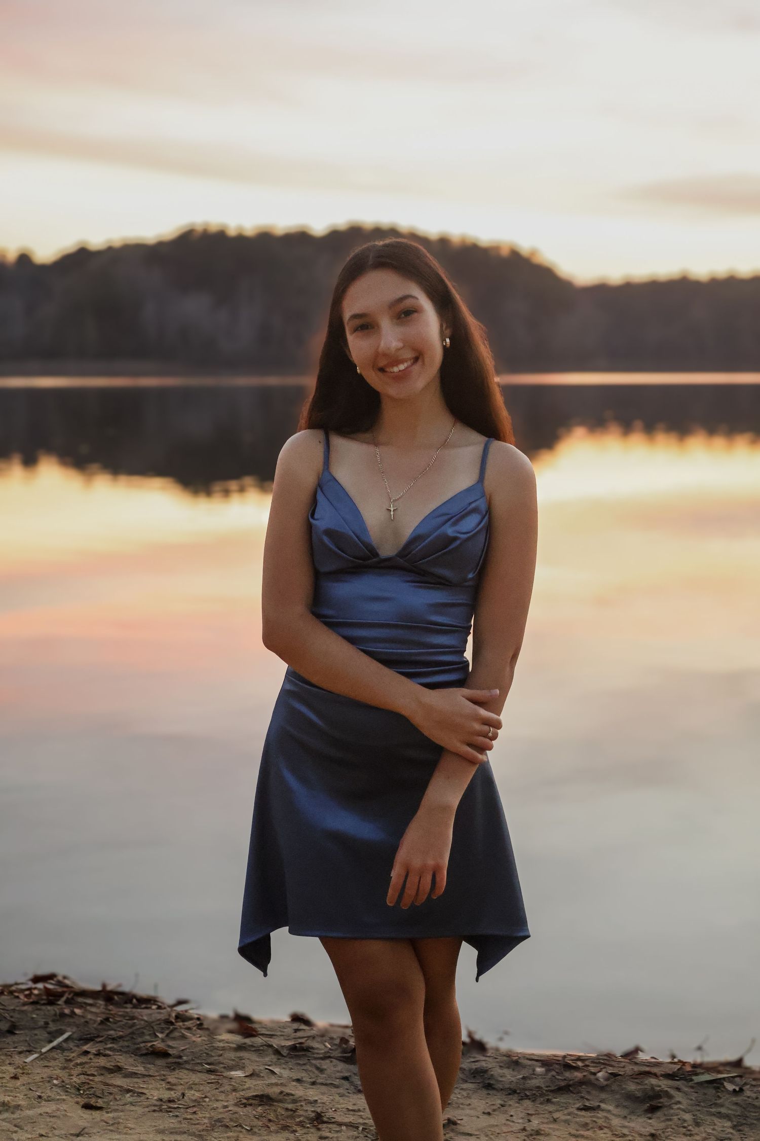 Smiling young woman in blue satin dress posing by reflective lake at colorful sunset.