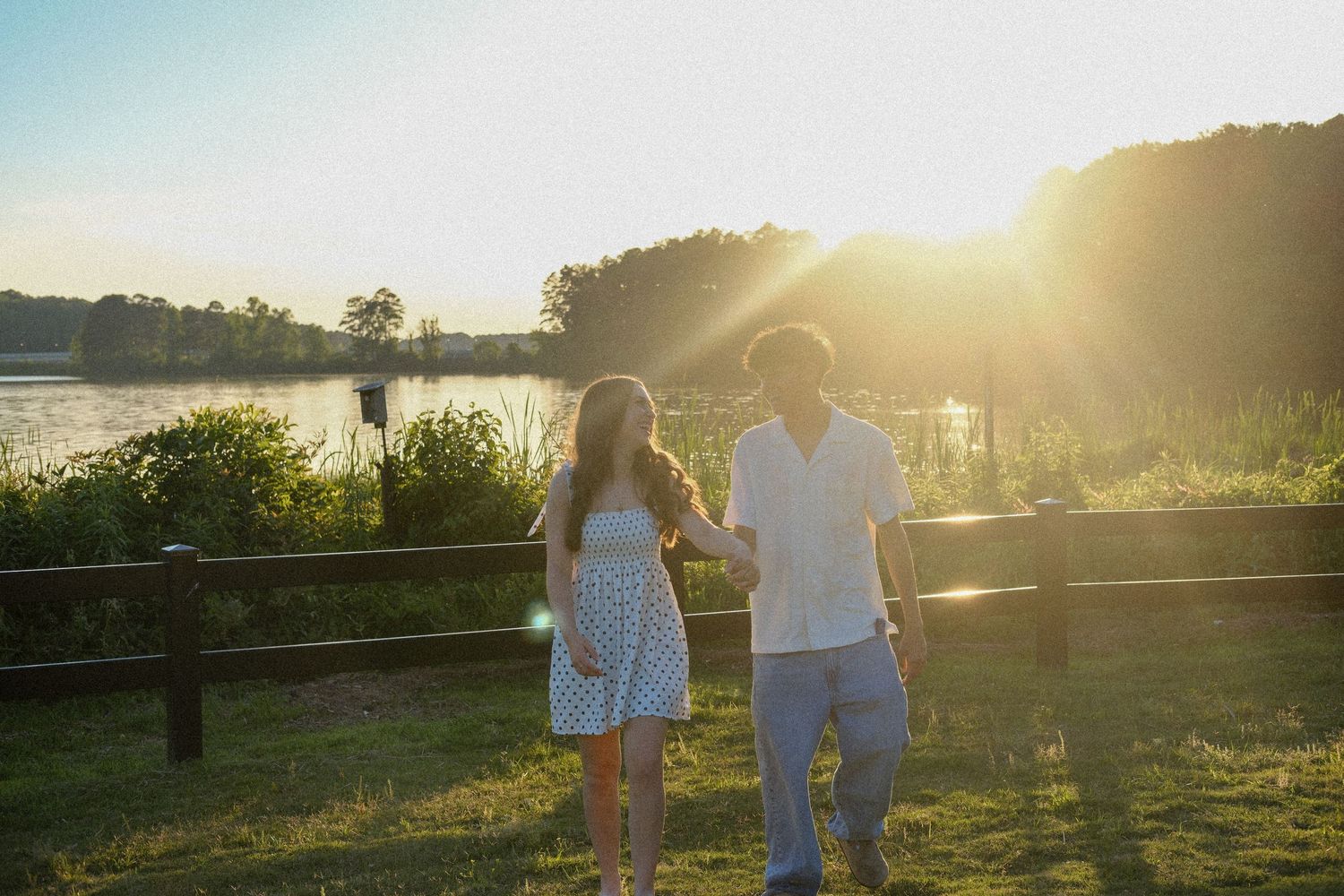 Couple holding hands at sunset by a lakeside fence surrounded by lush greenery in warm golden light.