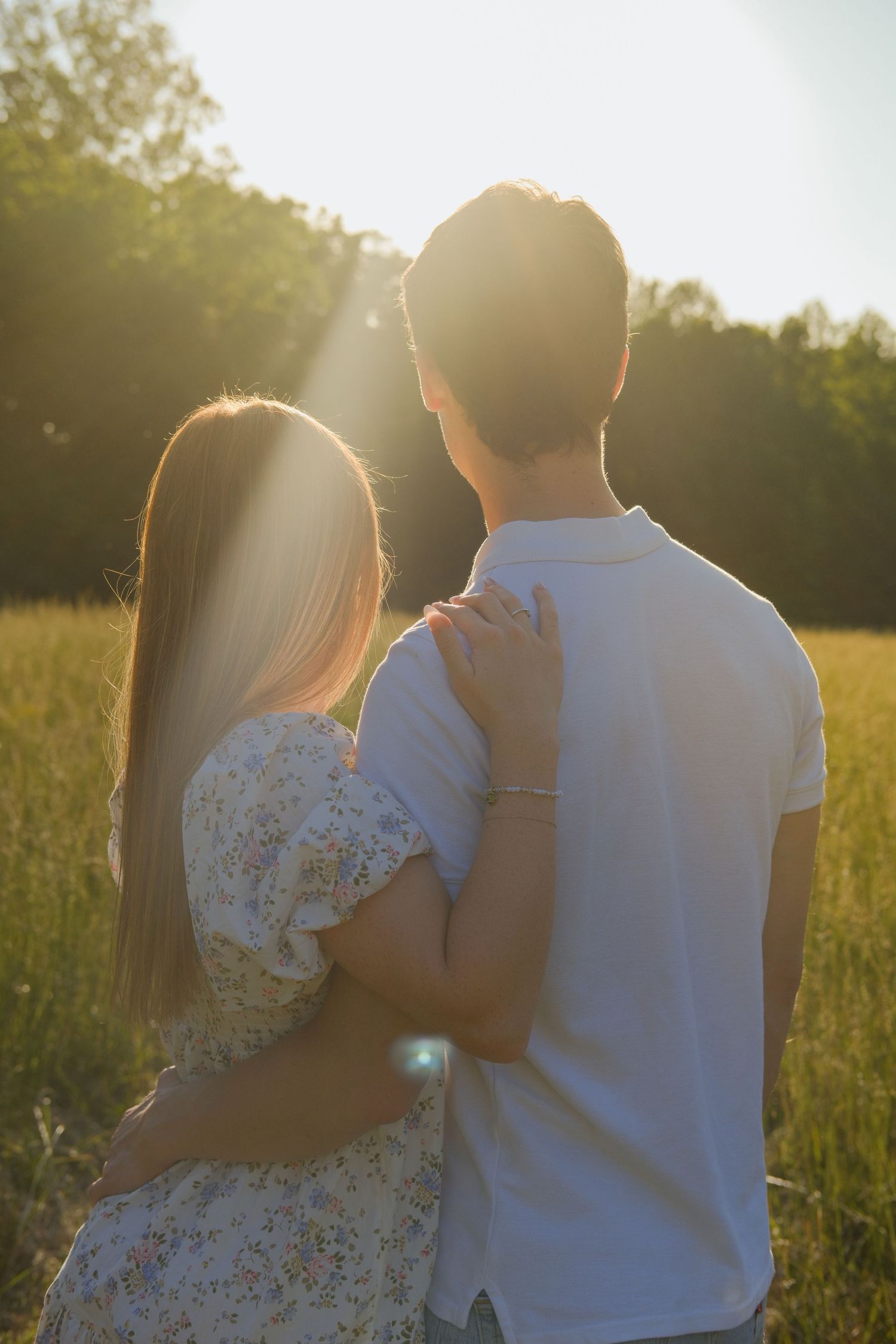 A couple embraces in a sunlit field at golden hour, viewed from behind with warm light filtering through trees.