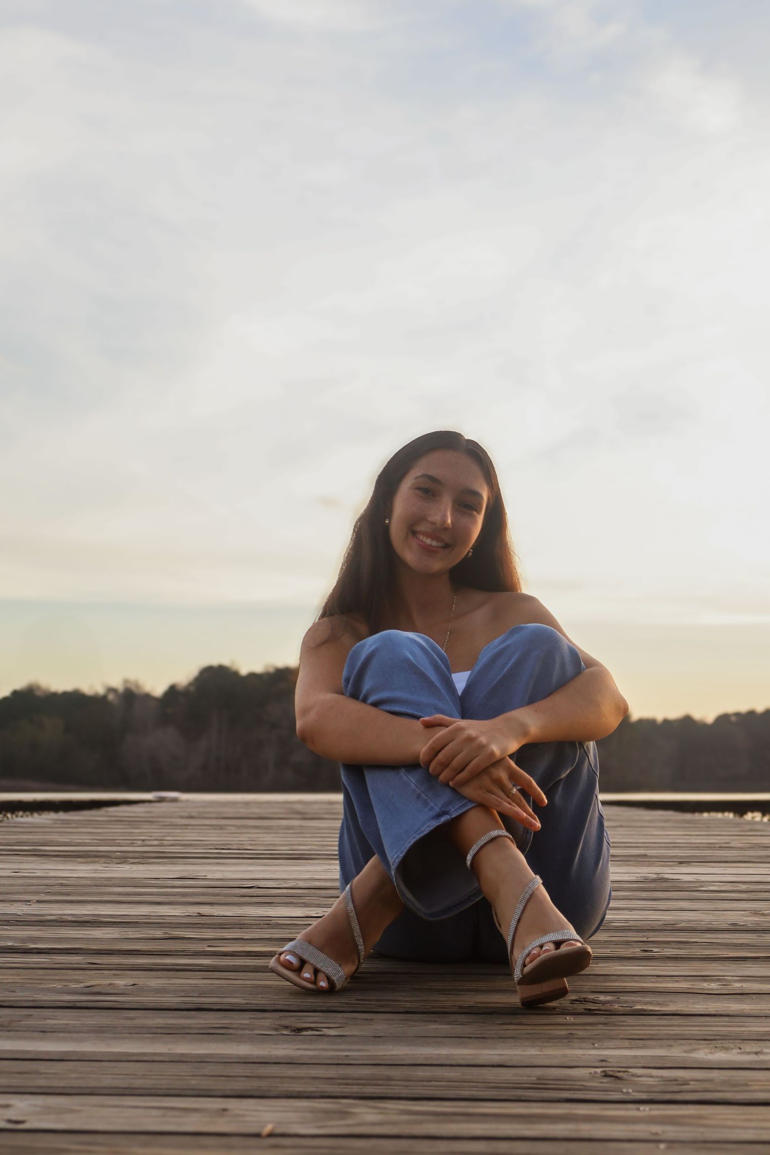 Young woman in blue outfit sitting on wooden dock at sunset, smiling at camera with knees drawn up.