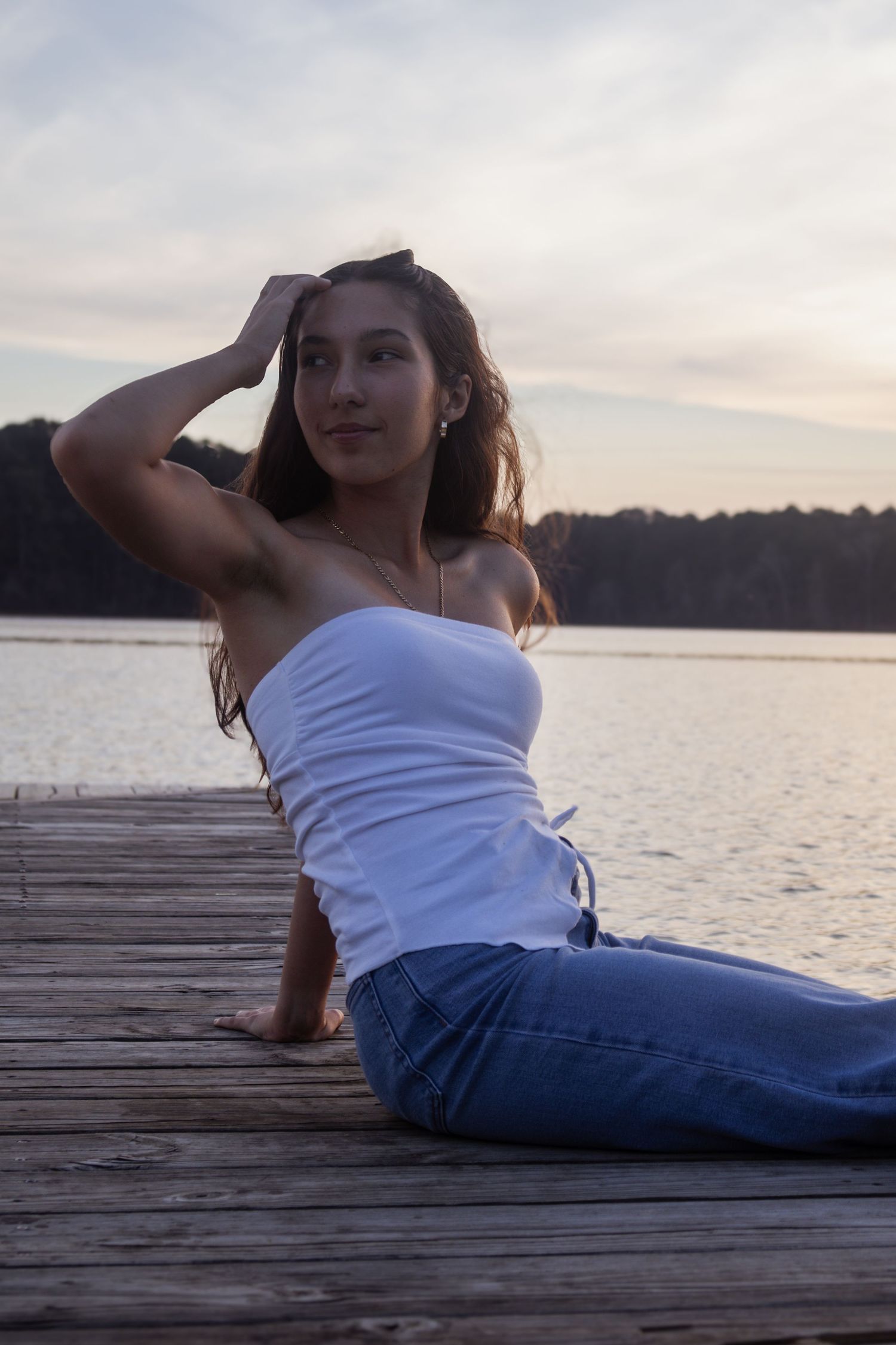 Young woman in white tube top and jeans sitting on wooden dock by lake at sunset.