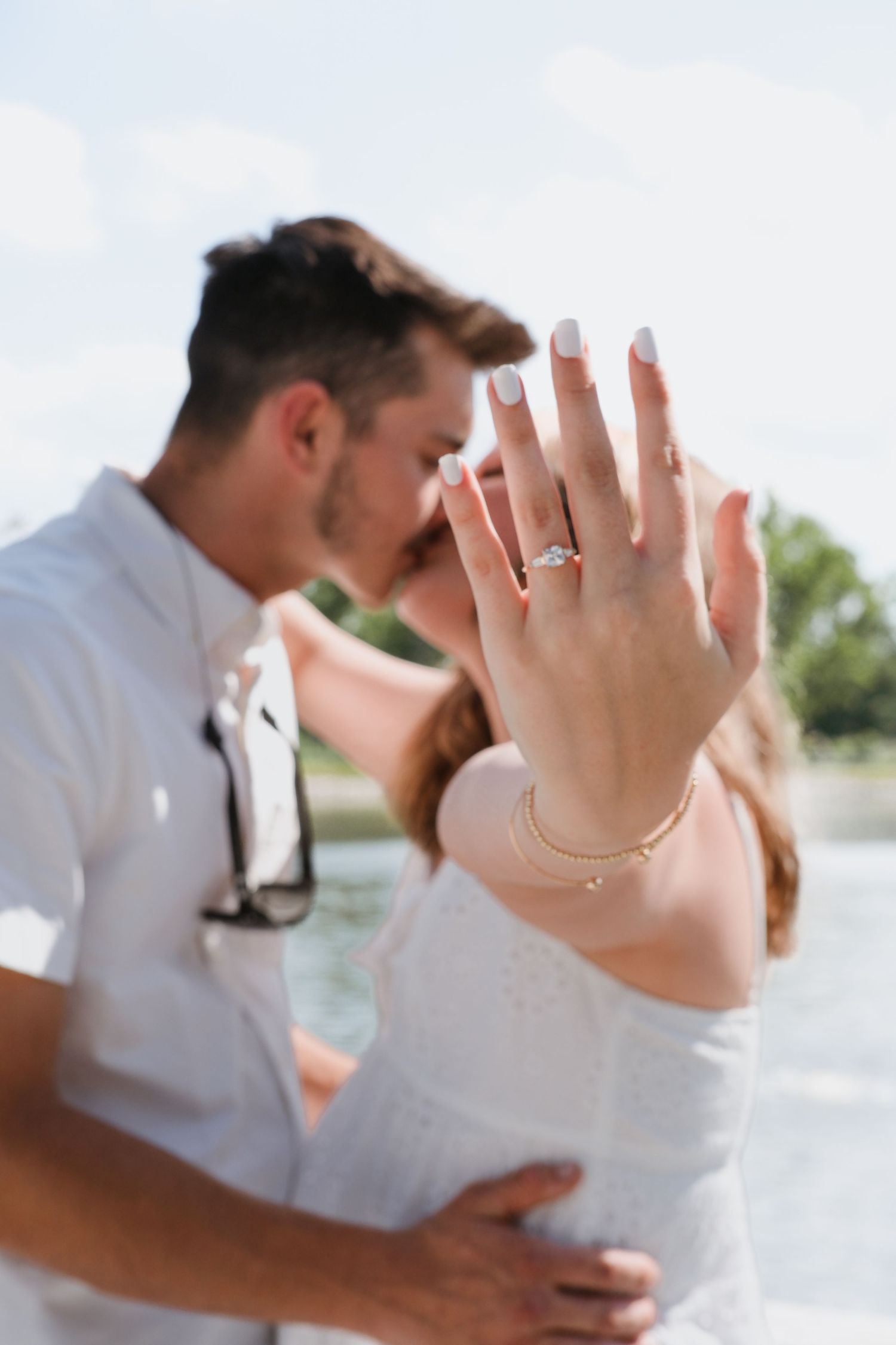 Couple kissing outdoors while showing off a diamond engagement ring with manicured nails near a lake.
