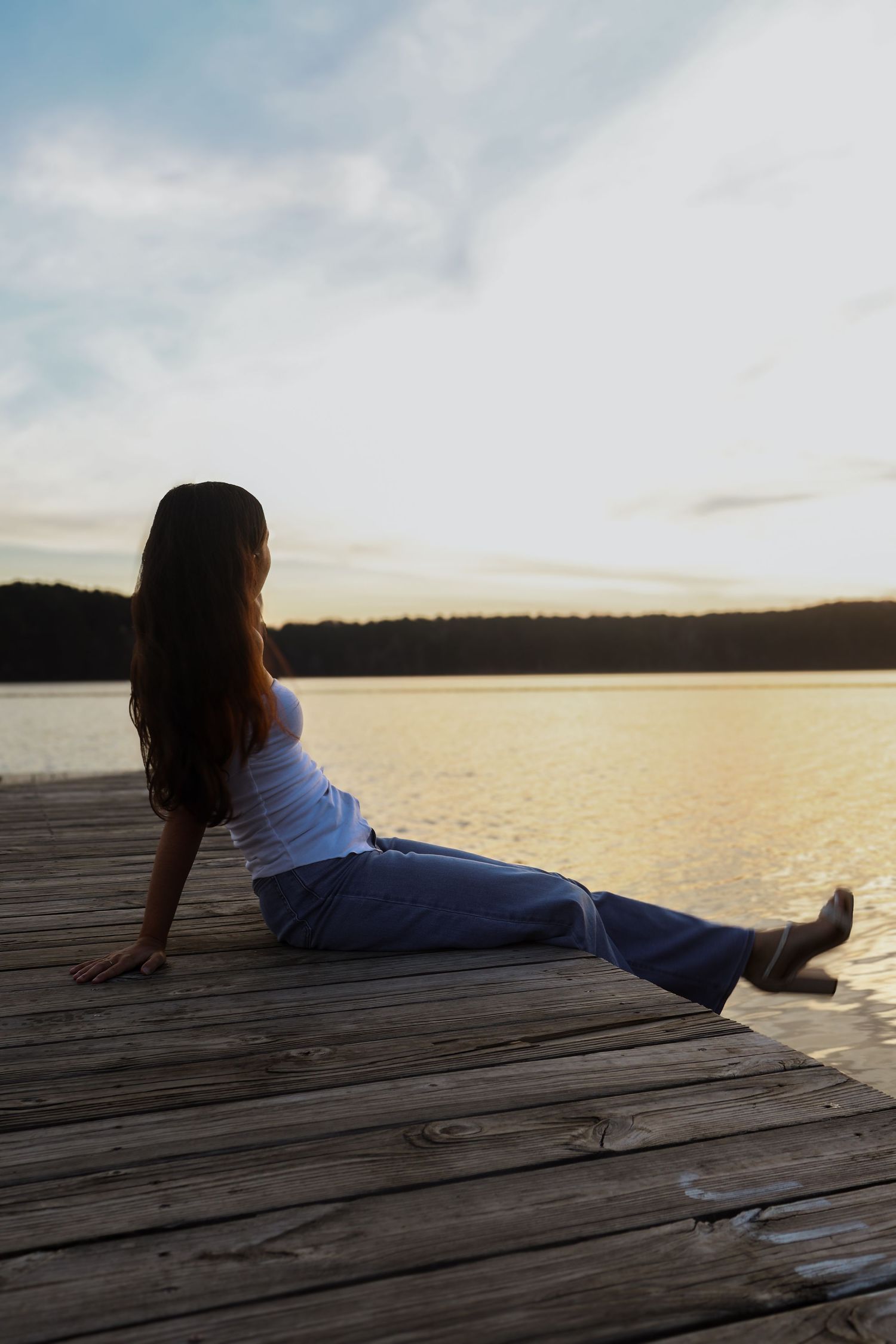 Young woman in white top and jeans sitting on wooden dock gazing at calm lake during golden sunset.