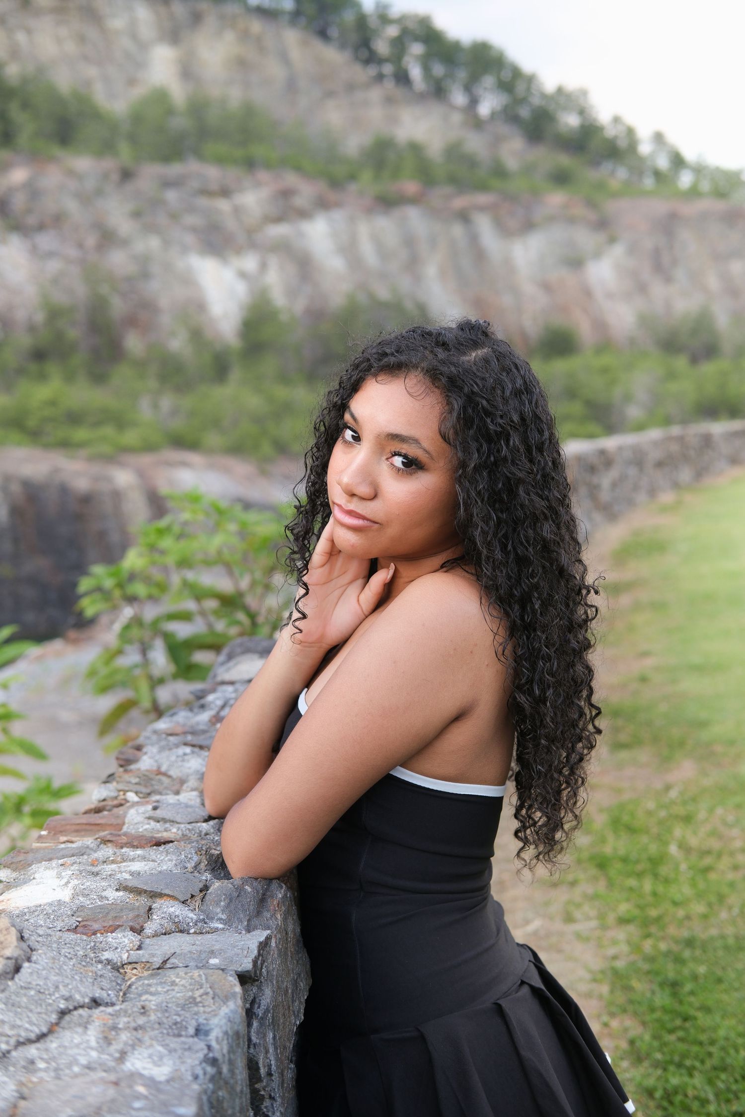 Young woman with long curly black hair in a black strapless dress, posing by a stone wall with rocky cliffs behind her.