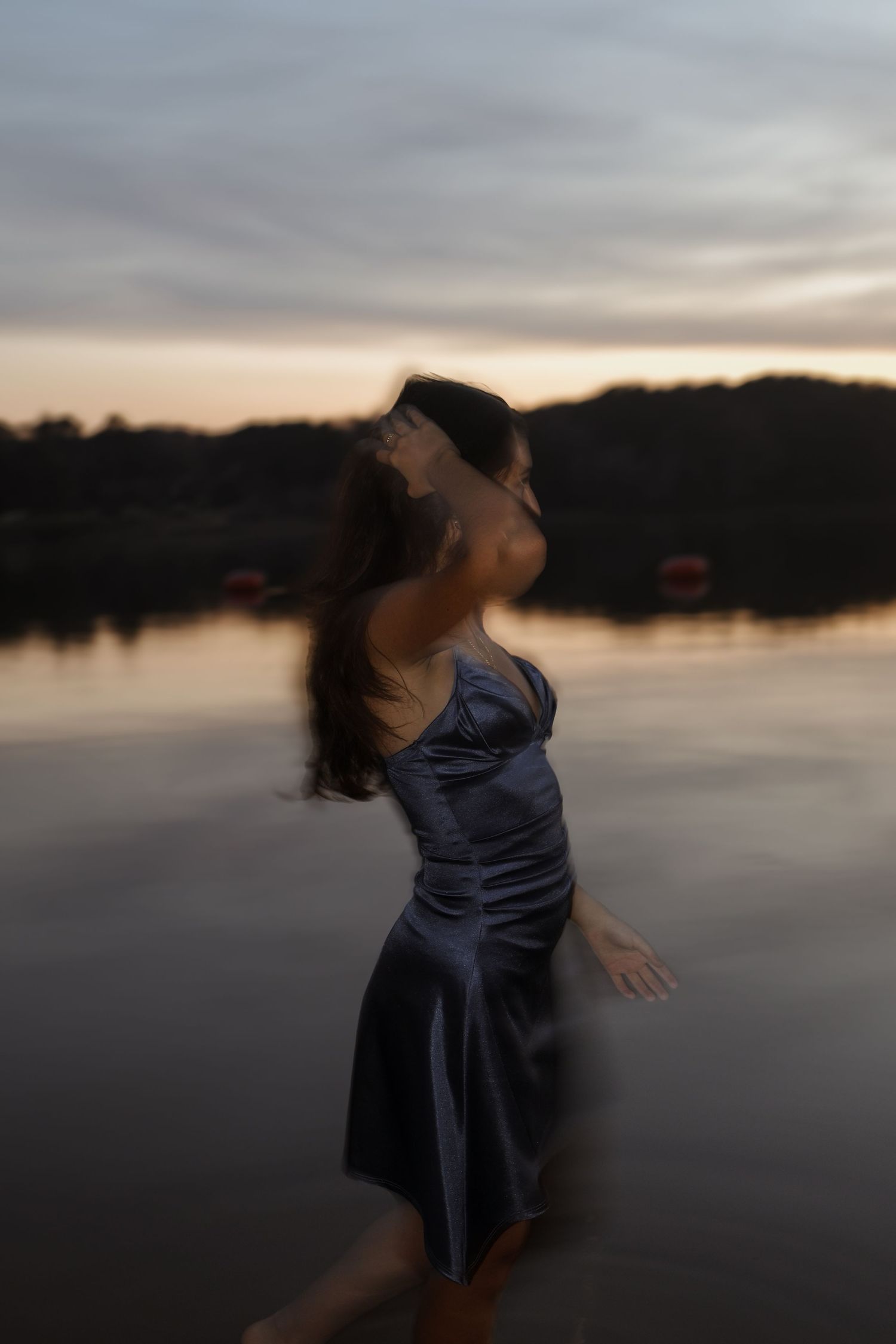 Woman in silver satin dress dancing gracefully by a lake at dusk with motion blur effect.