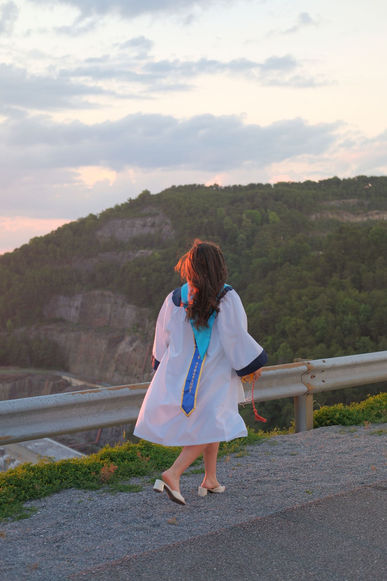A graduate in white cap and gown stands at a scenic overlook, gazing at lush green cliffs at sunset.