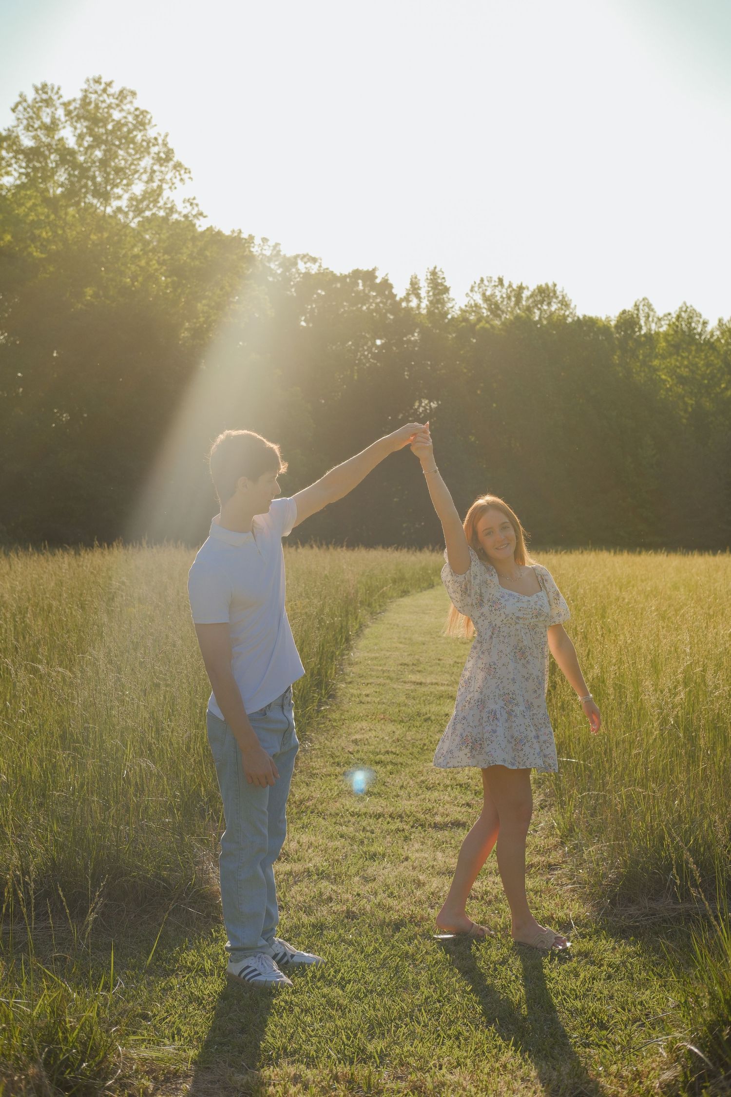 Couple dancing in a sunlit field, man spinning woman in floral dress, golden hour light with trees in background.