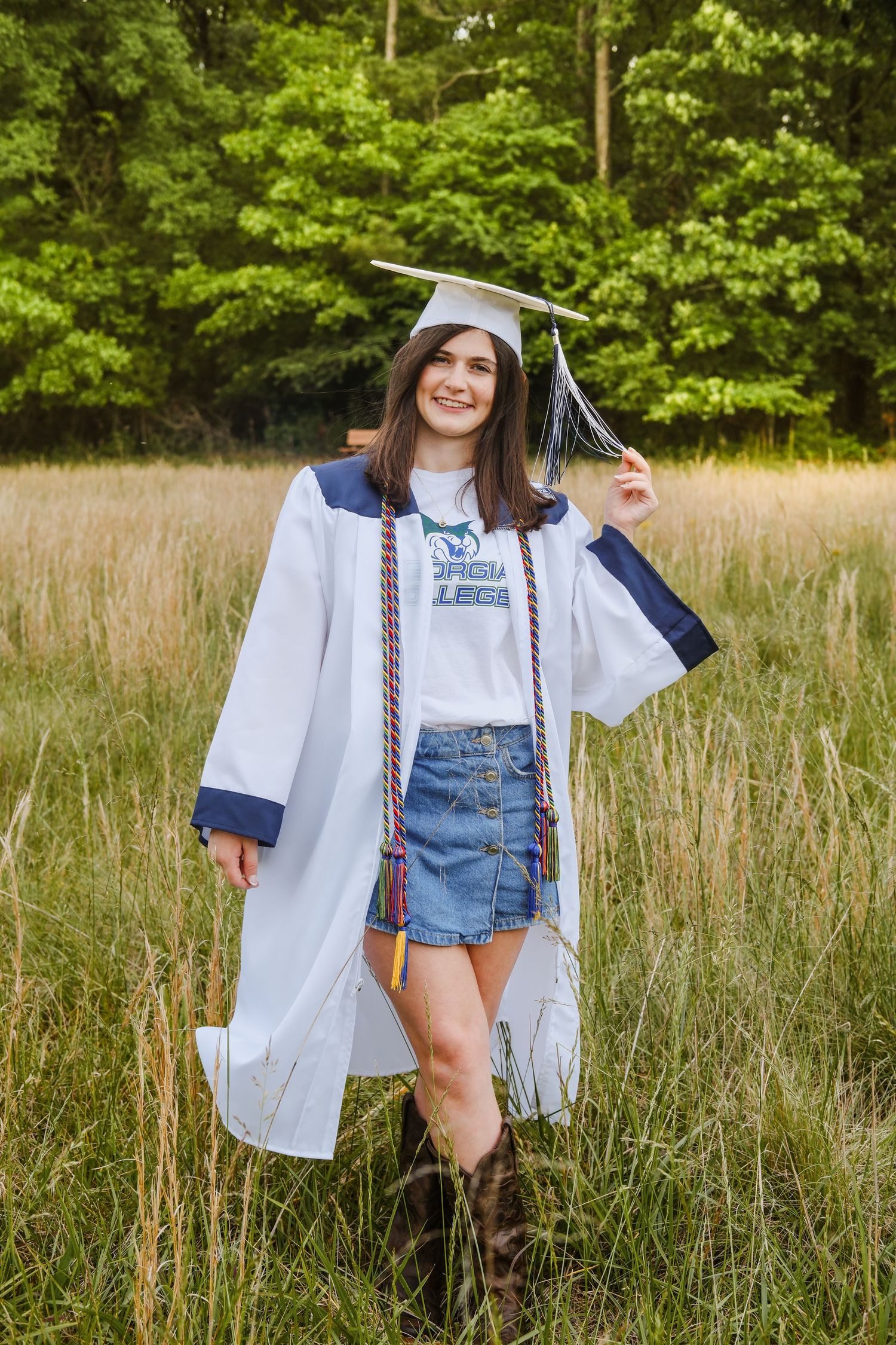 Graduate in white cap and gown with honor cords posing in a golden meadow.