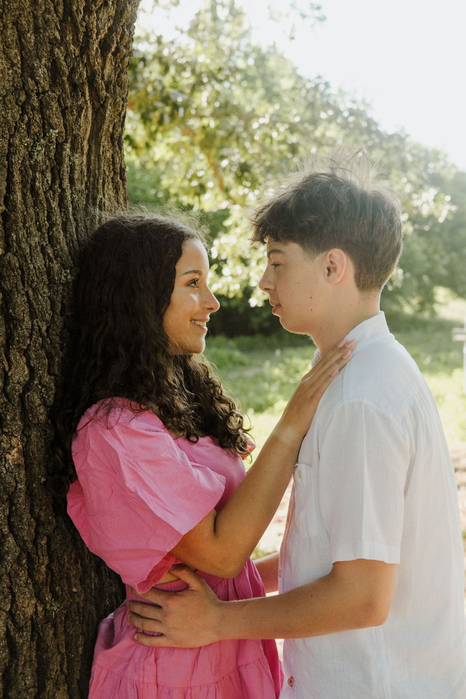 Young couple sharing a romantic moment leaning against a tree trunk in a sunny outdoor park setting.