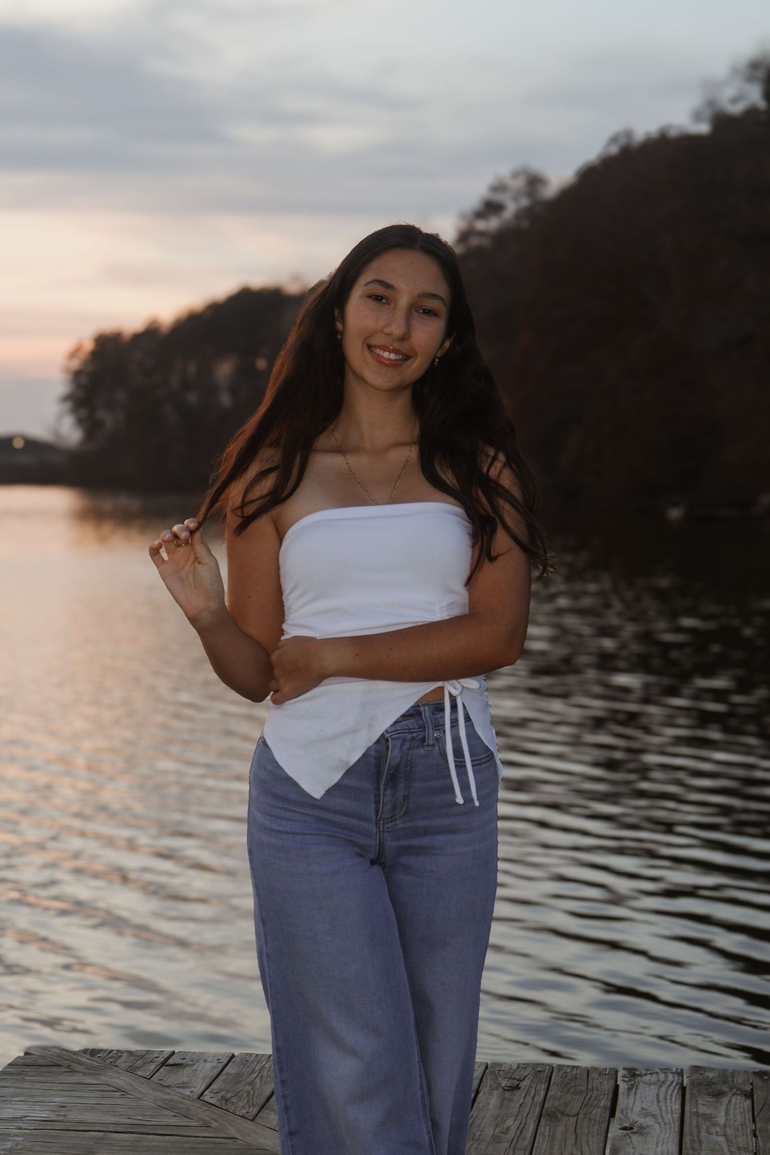Young woman in white tube top and jeans smiling on a dock at sunset with a lake and trees behind her.