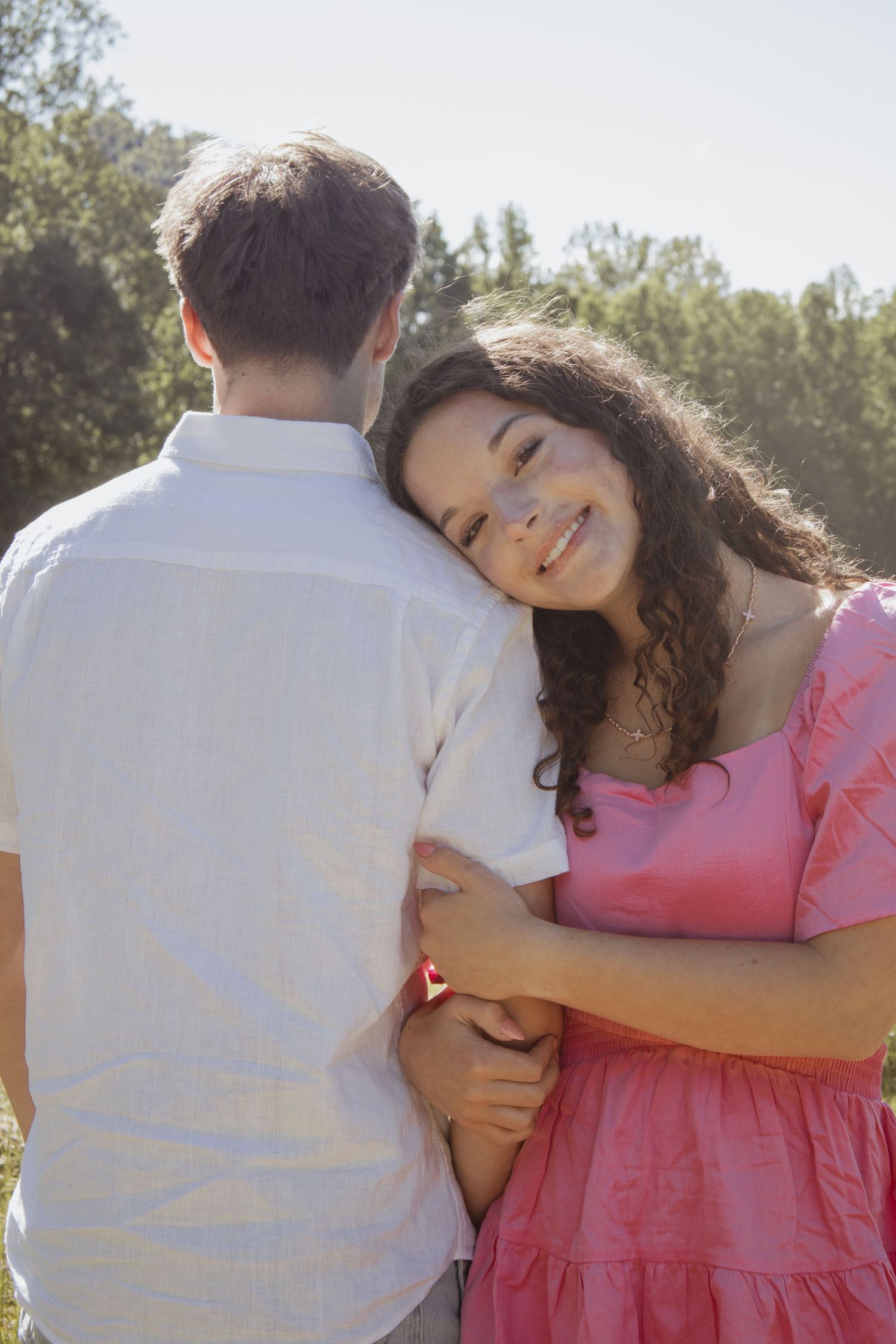 Young woman in pink dress smiling while hugging a man in white shirt outdoors in sunny field.