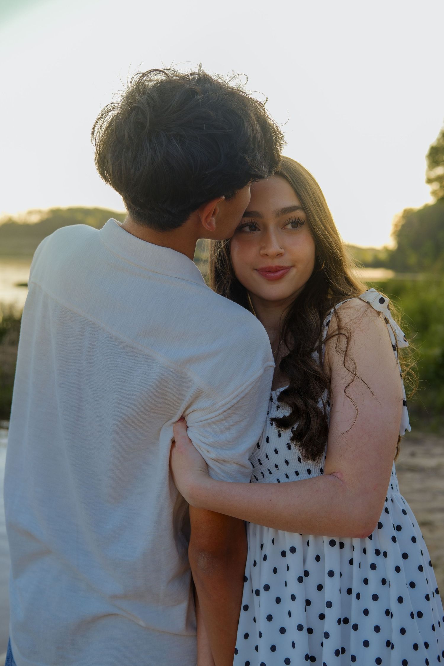 A young couple embraces outdoors at sunset, the smiling woman in a polka dot dress looking back at the camera.