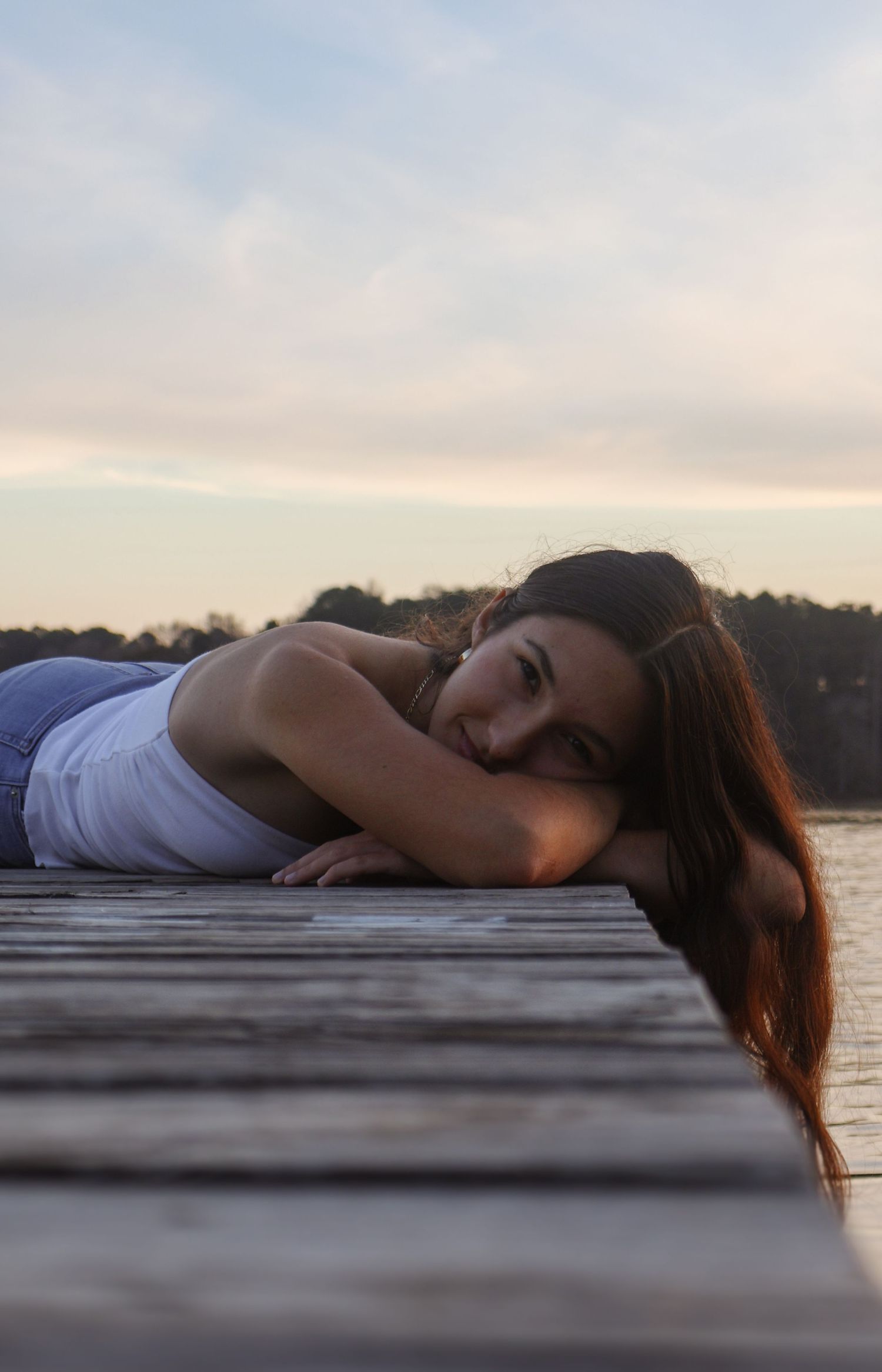 Young woman with long brown hair lying on a wooden dock by a lake at sunset, gazing at the camera.