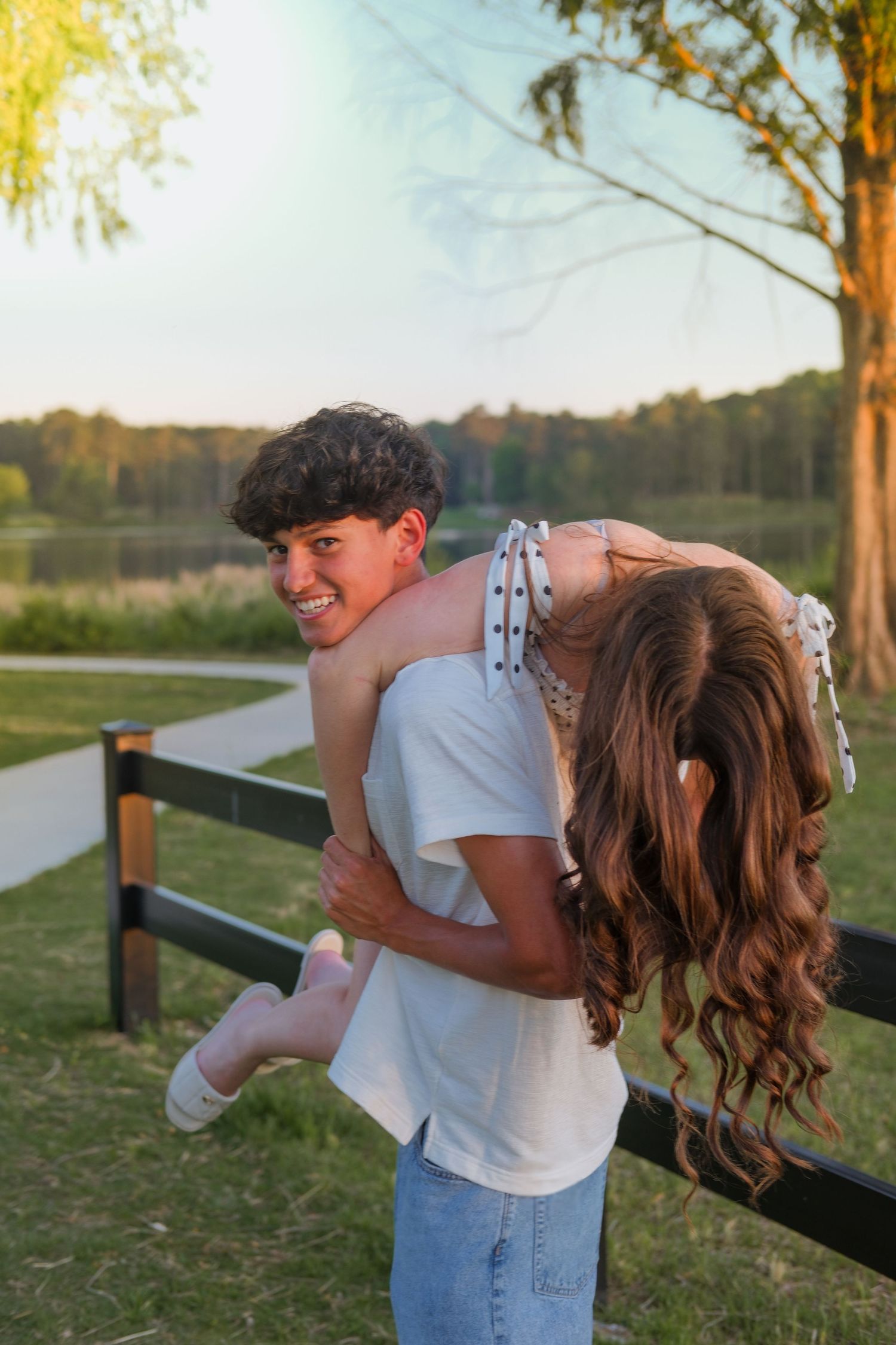 Young couple at golden hour, boy lifting girl near wooden fence by a lake surrounded by trees.