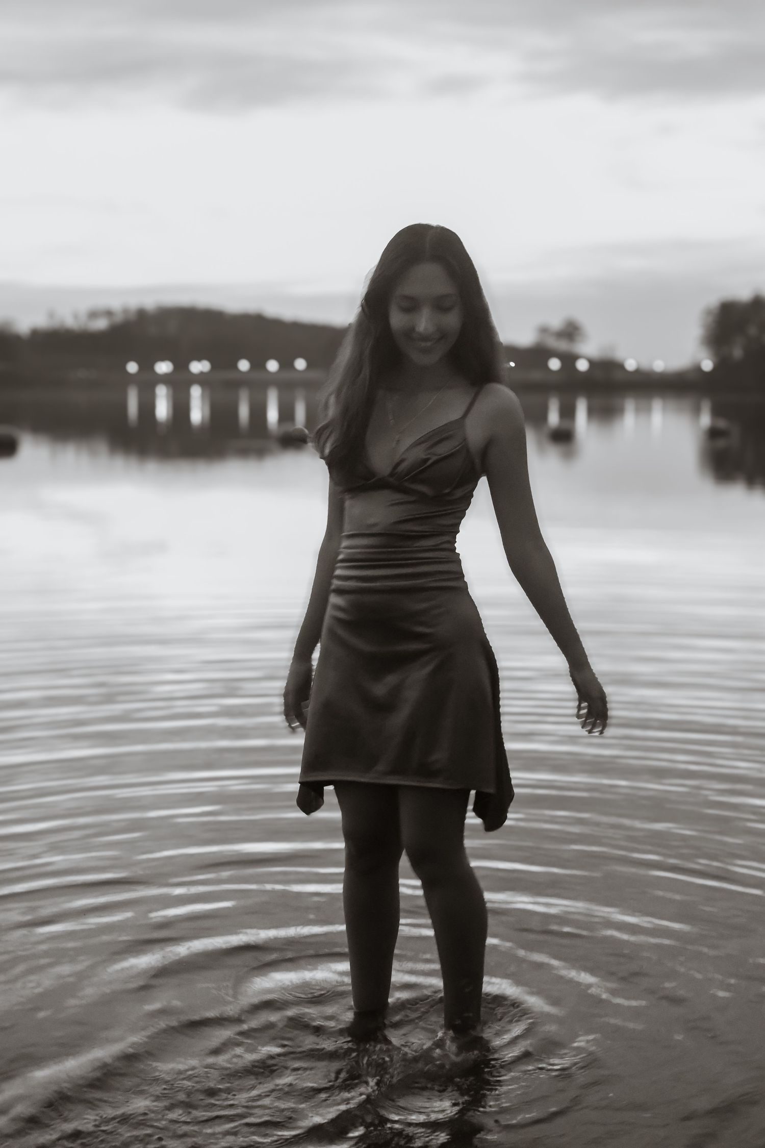 Young woman in a dark dress standing in shallow water at dusk, smiling, with a bridge and trees in the background.
