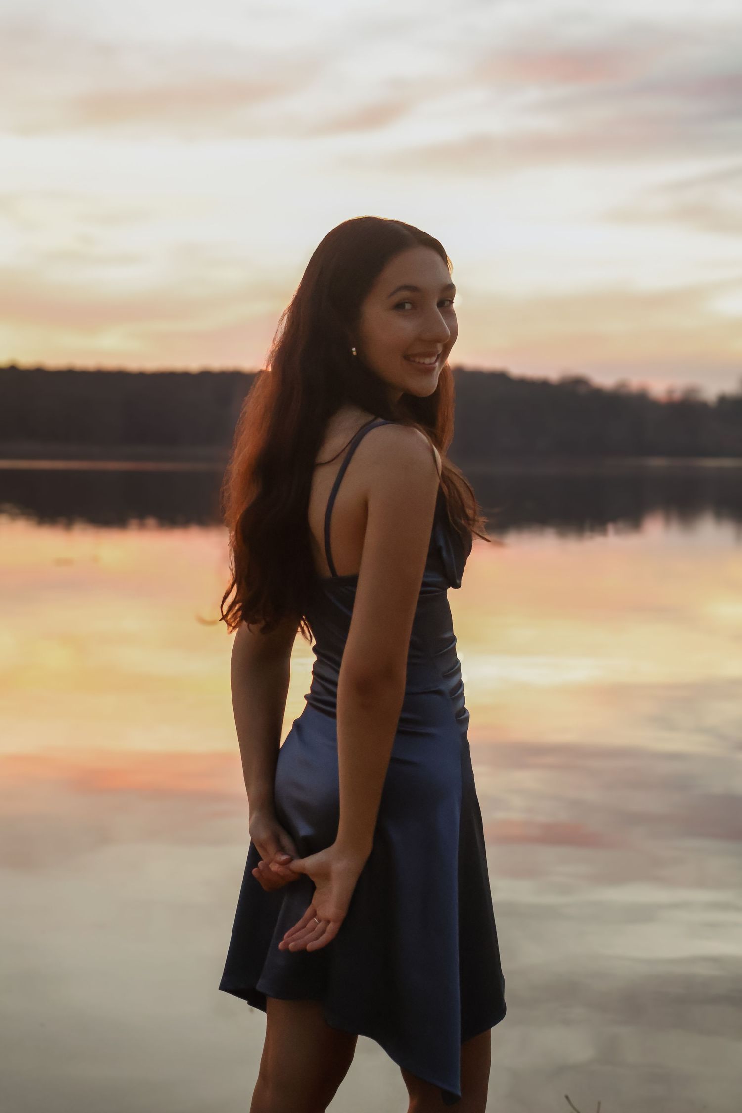 Young woman in a blue dress smiling over her shoulder by a reflective lake during a colorful sunset.