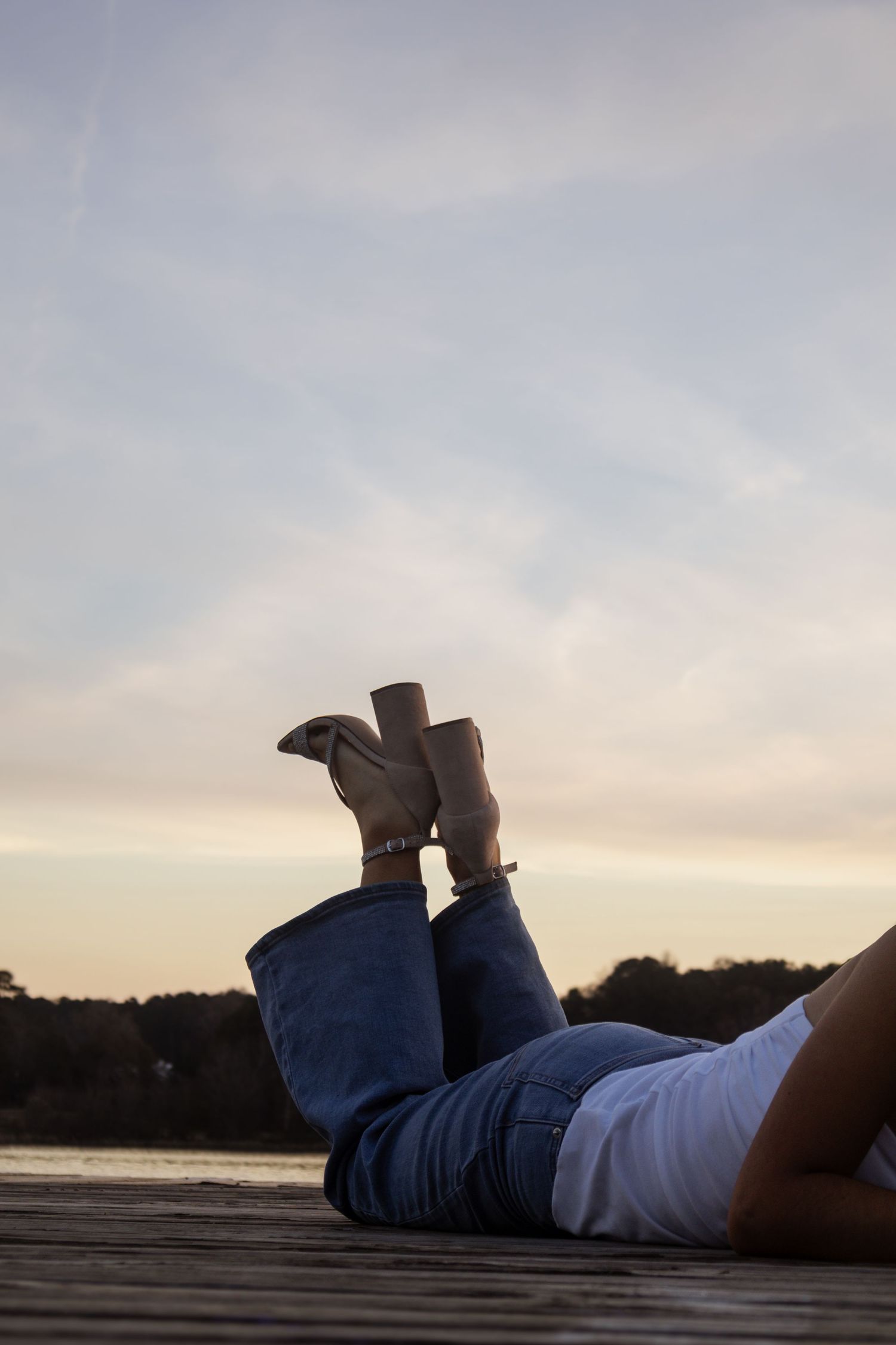 Person lying on wooden dock in jeans and boots, legs raised against a serene sunset sky near a lake.