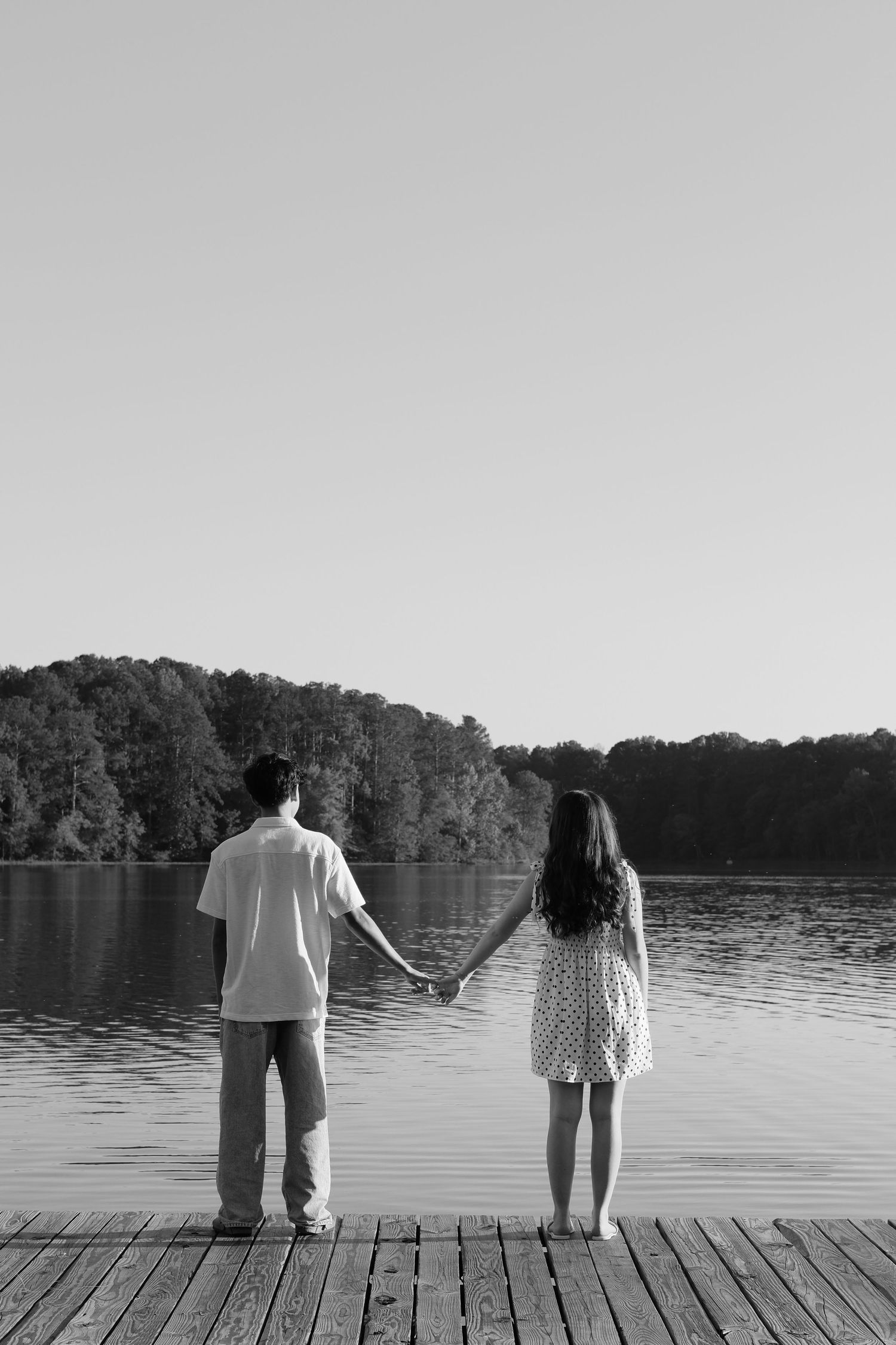 Black and white photo of a couple holding hands on a wooden dock overlooking a calm lake with trees.