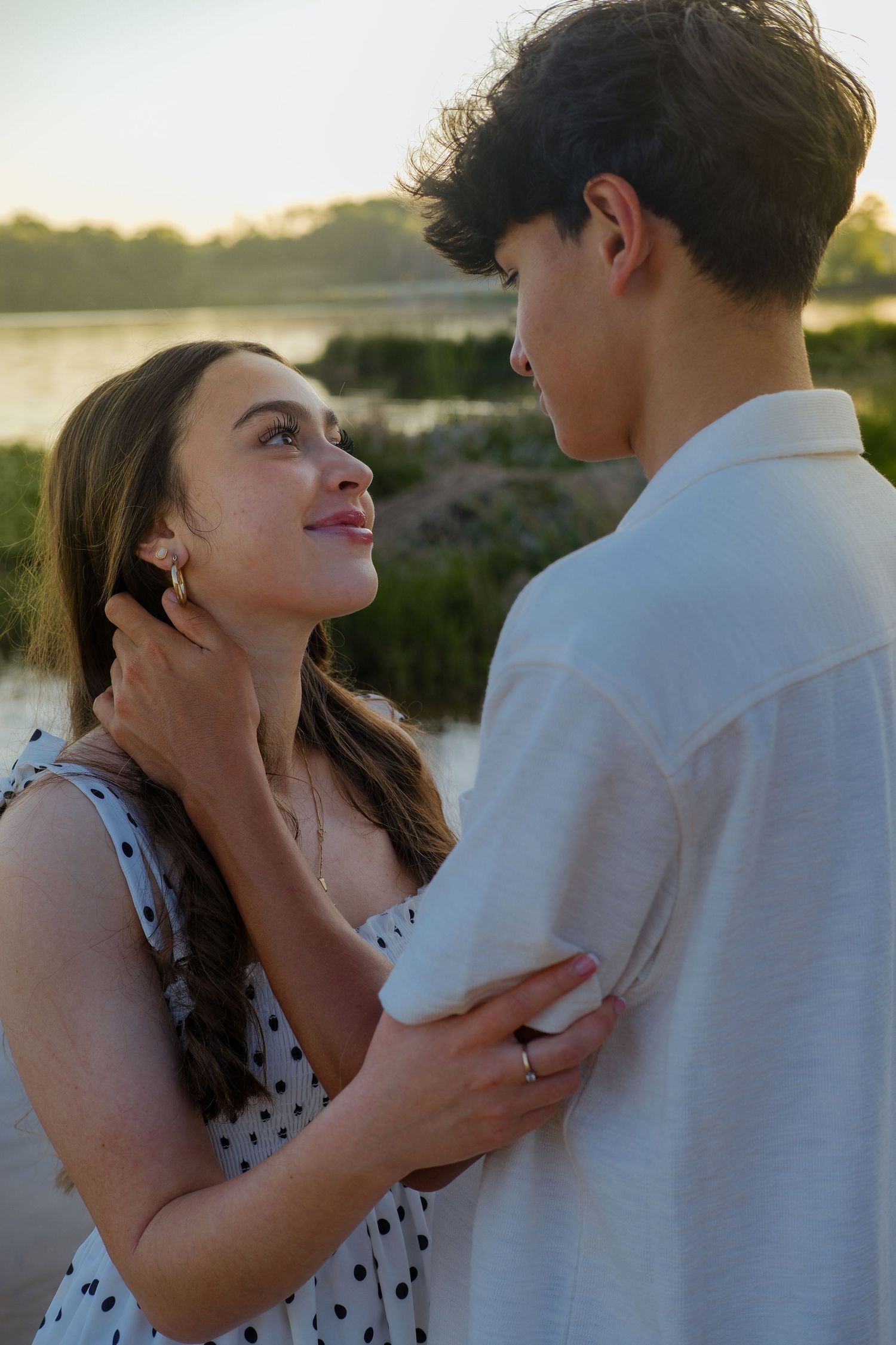 Young couple sharing a romantic moment outdoors near a lake at sunset, woman in polka dot dress gazing up at man.