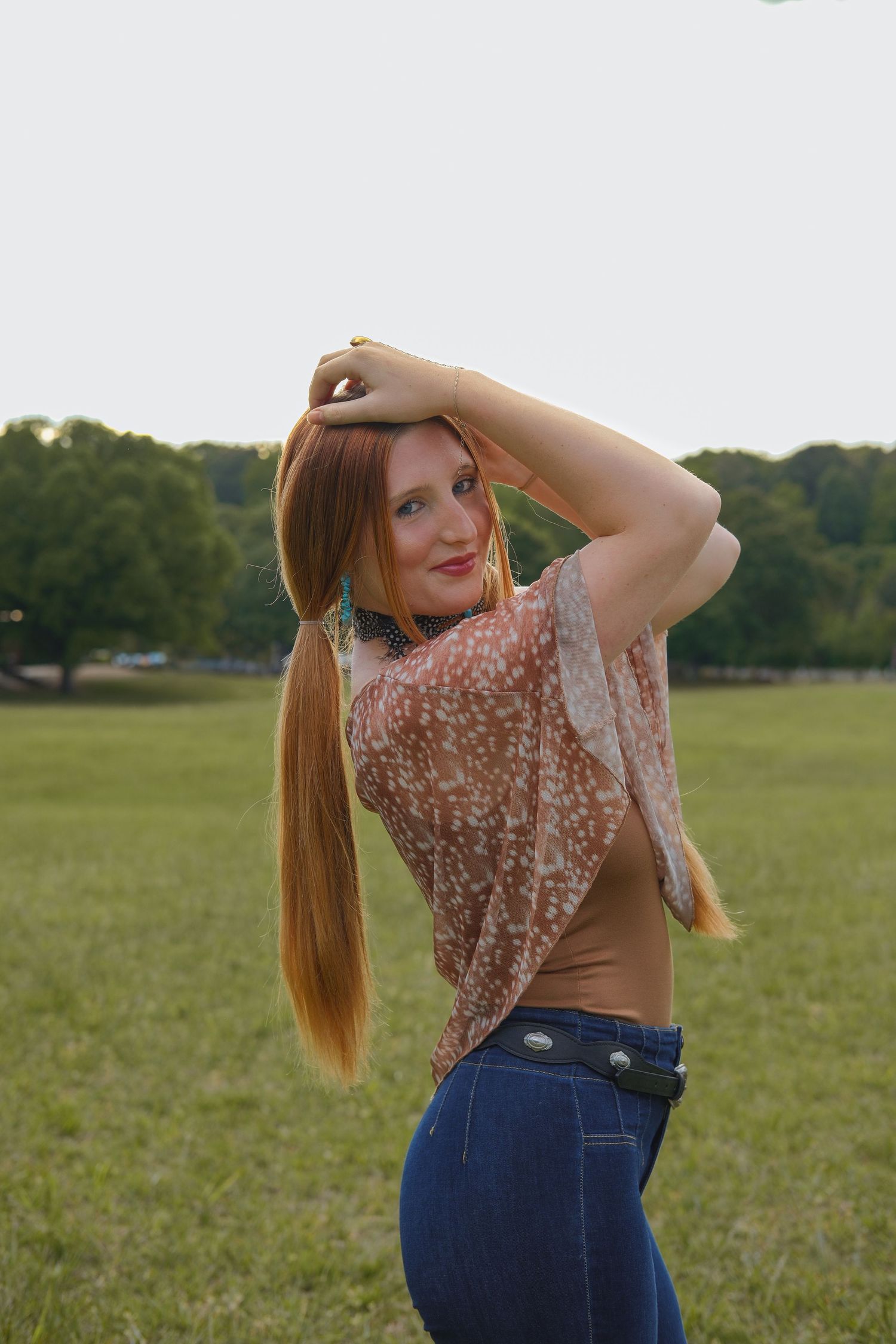 Redhead woman in floral blouse and jeans posing with hands in hair in a green park.