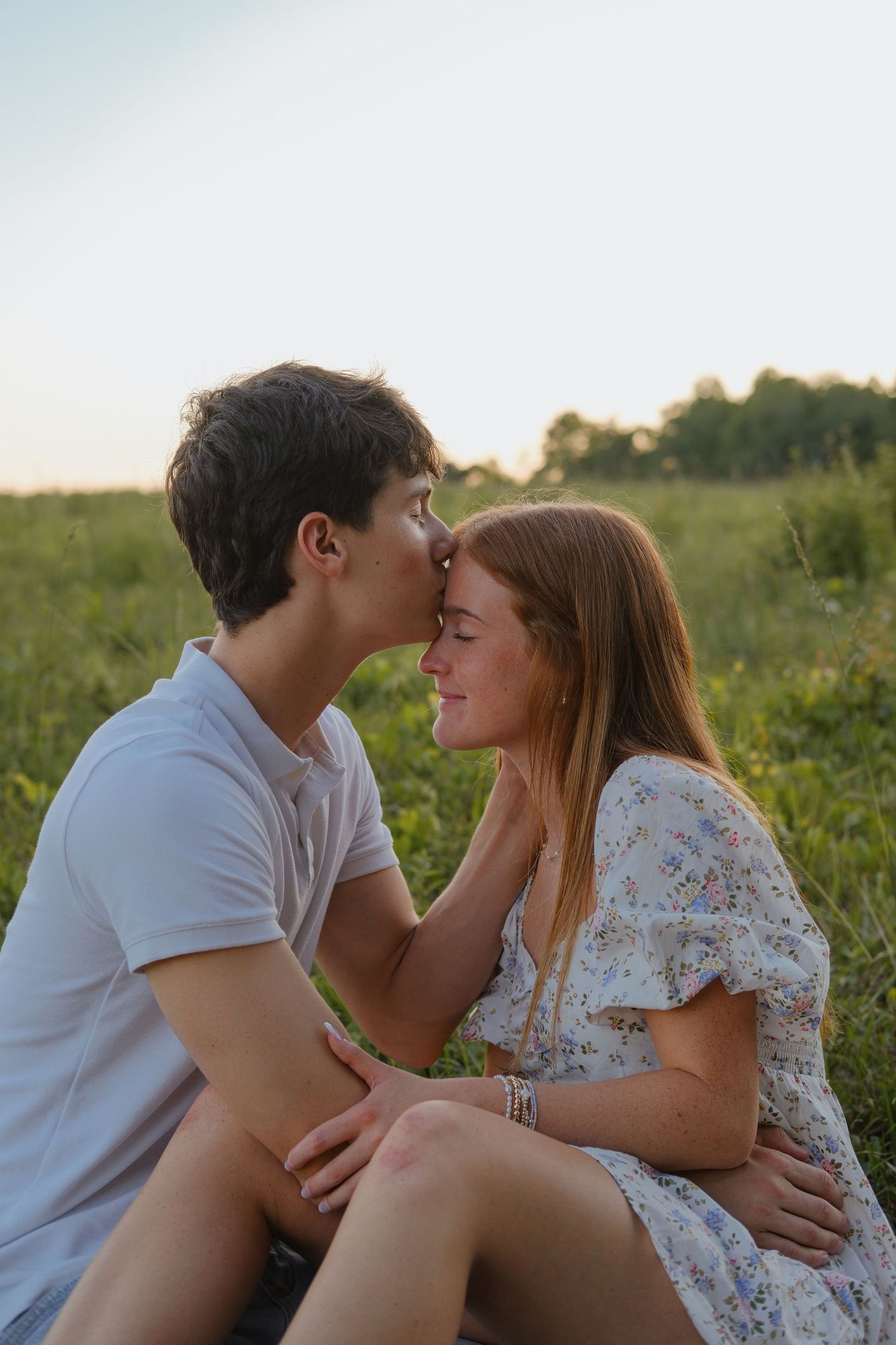 Young couple sharing a tender kiss in a green field at sunset, girl in floral dress smiling with eyes closed.