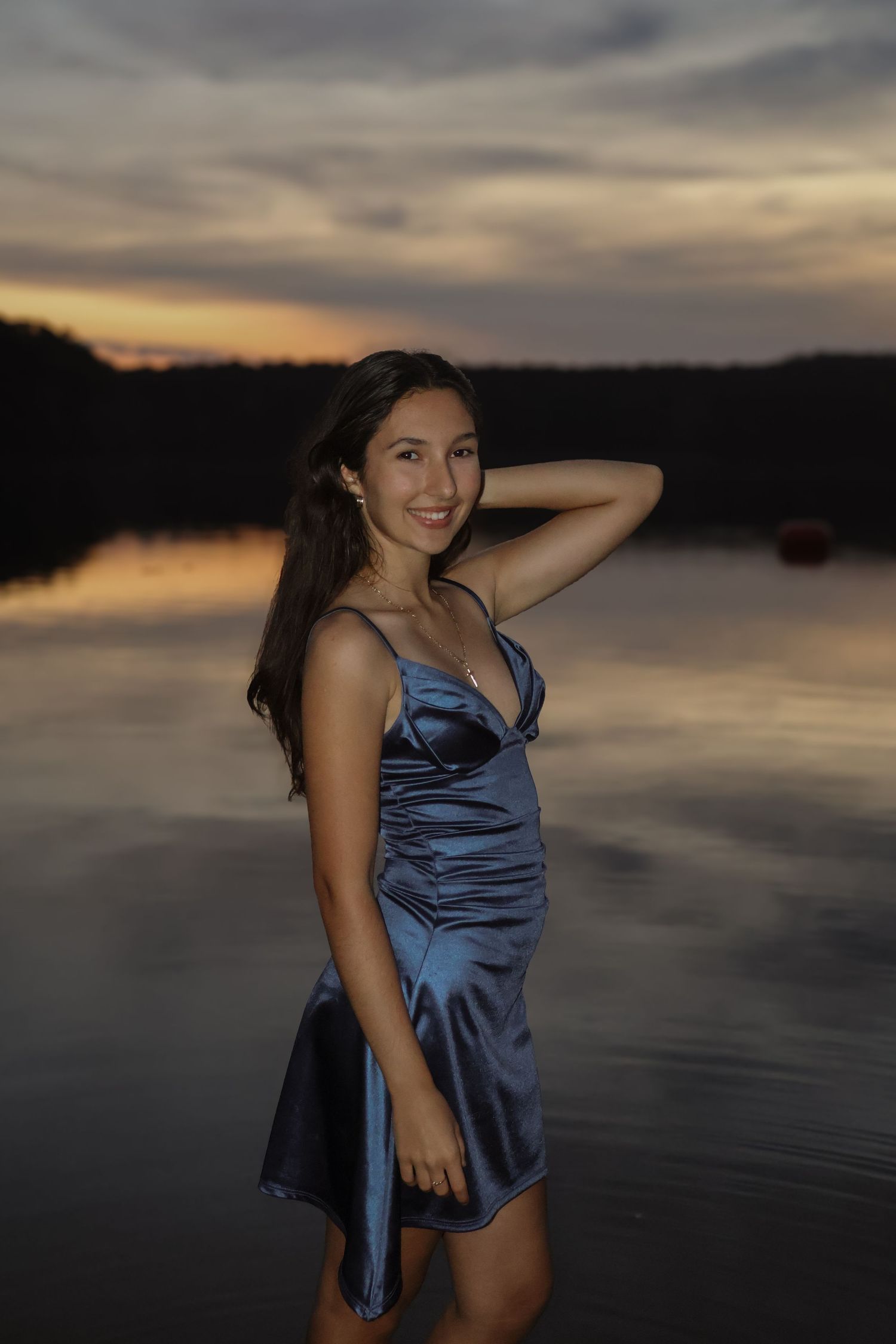 Young woman in blue satin dress smiling by reflective lake at sunset.