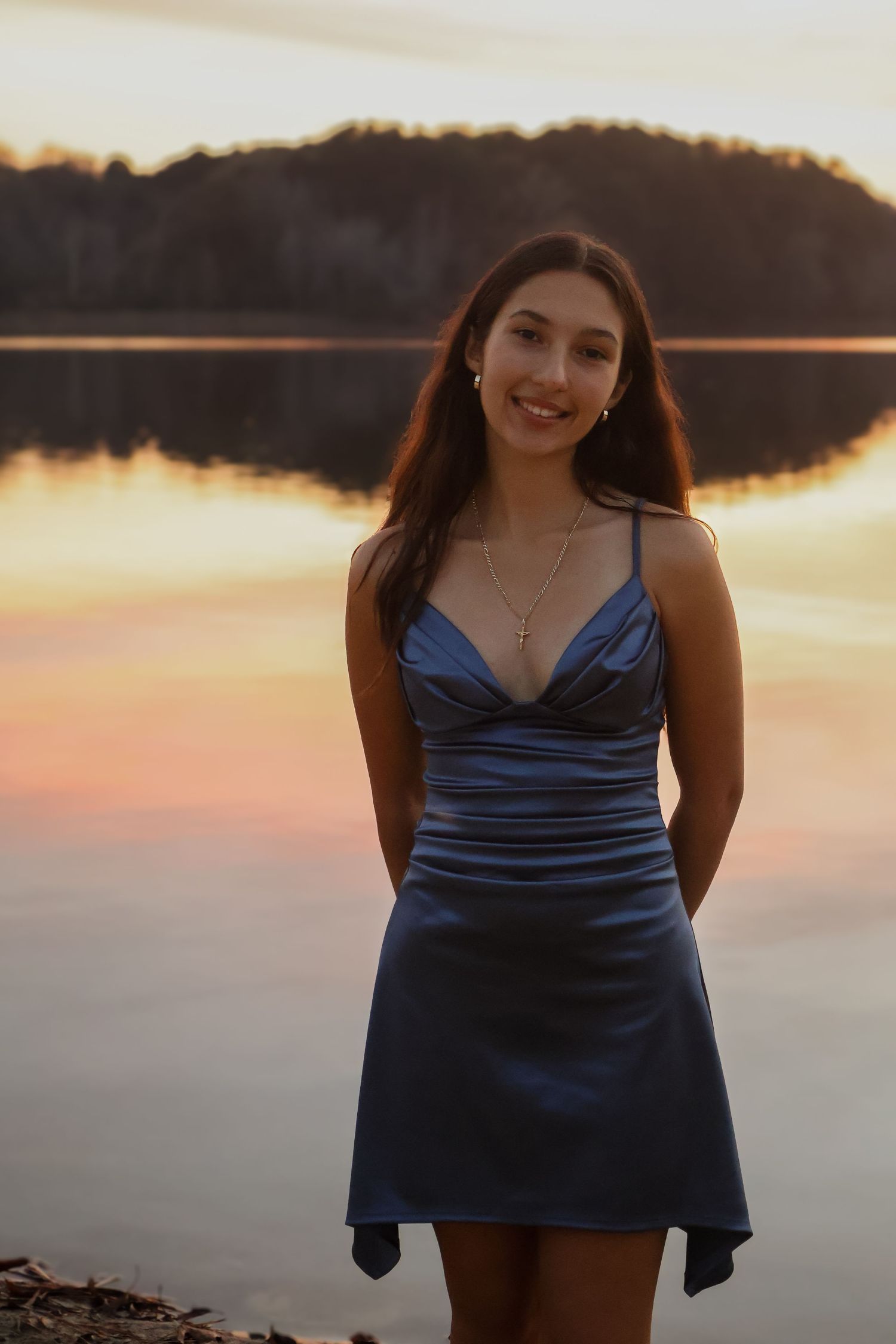 Young woman in blue satin dress smiling by a lake at sunset with golden reflections on the water.