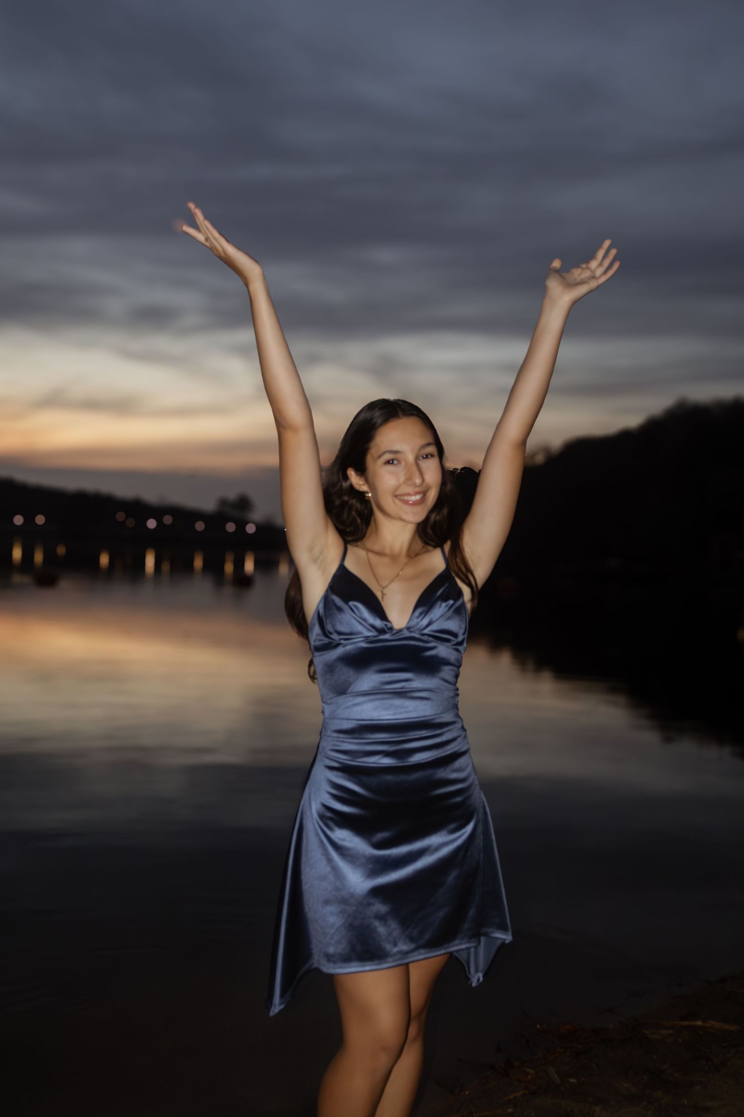 Young woman in blue satin dress celebrating with arms raised by a lake at dusk.