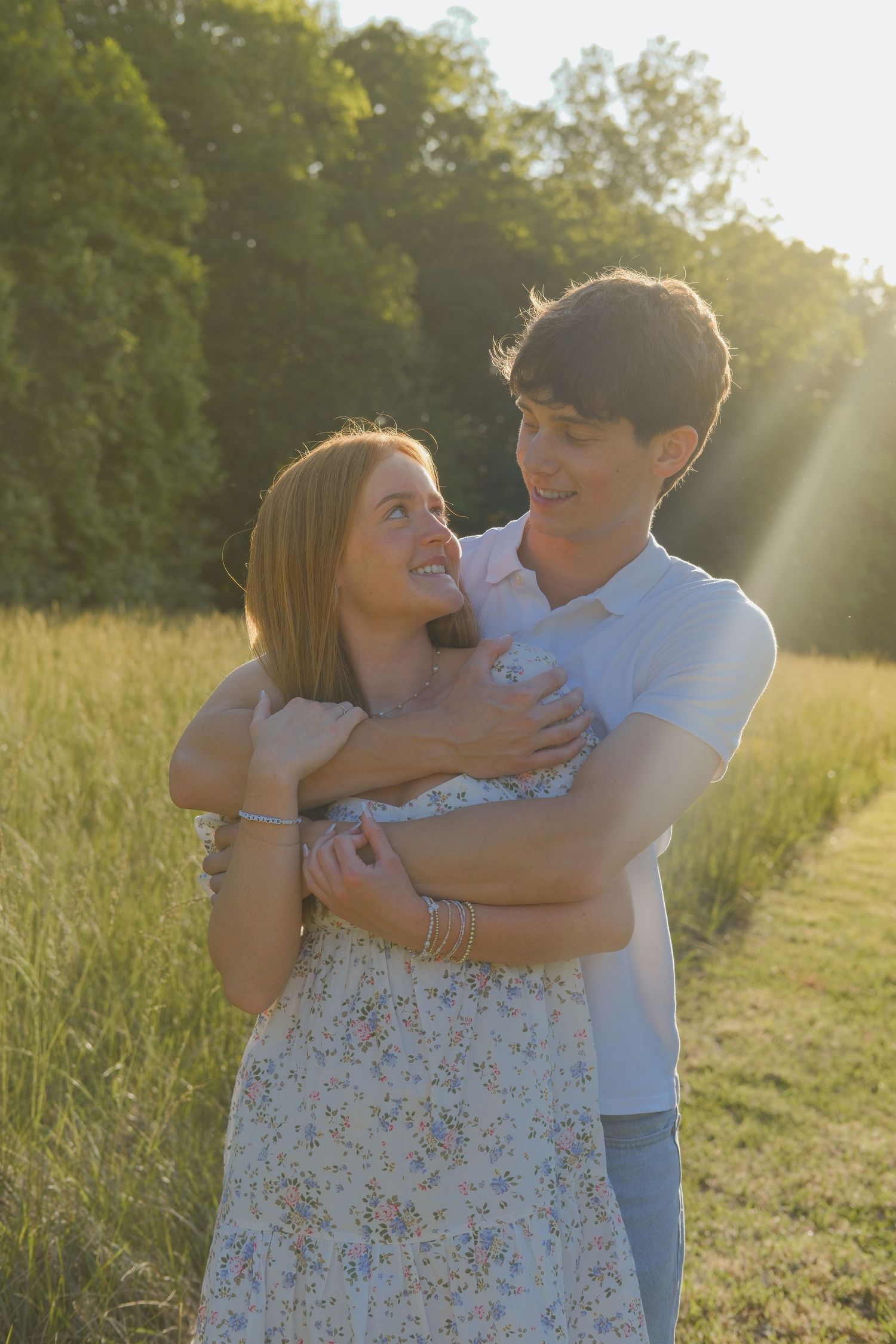 A young couple embraces in a sunlit field, smiling at each other, surrounded by lush green grass and trees.