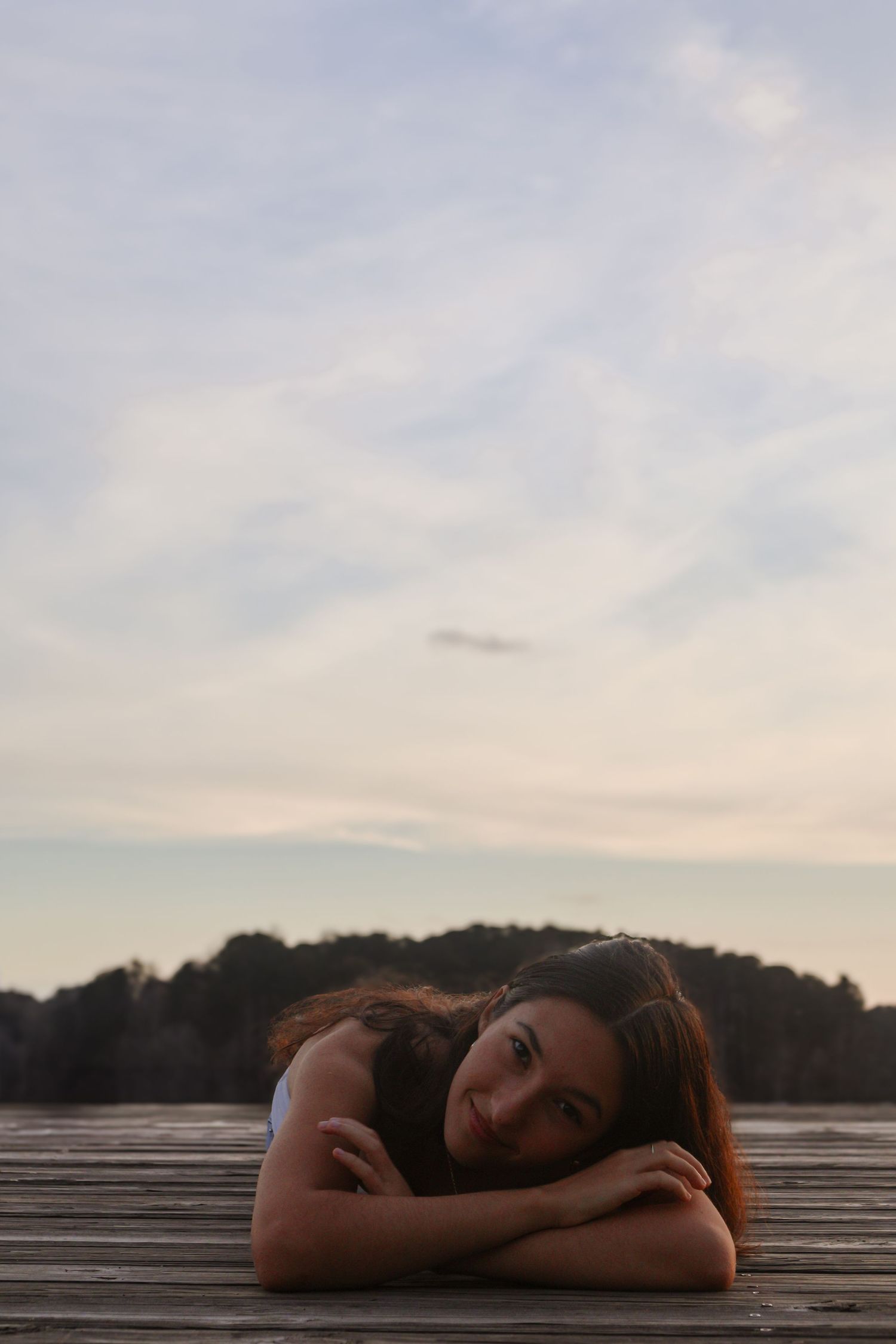 Young woman resting her chin on folded arms on a wooden dock at sunset with a serene lake and treeline in the background.