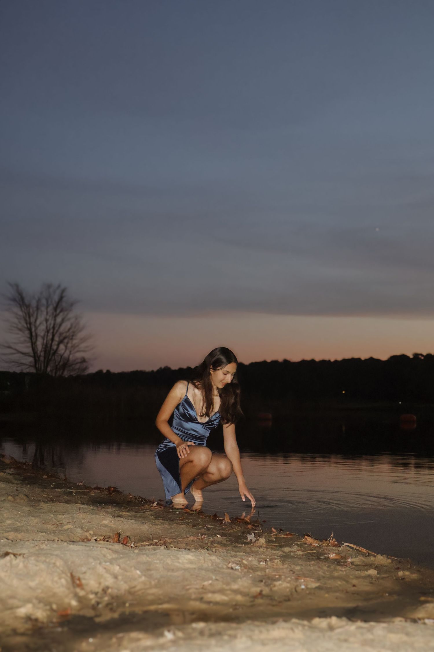 Young woman in blue dress crouching by a calm lake at dusk, touching the water's edge with a twilight sky behind her.
