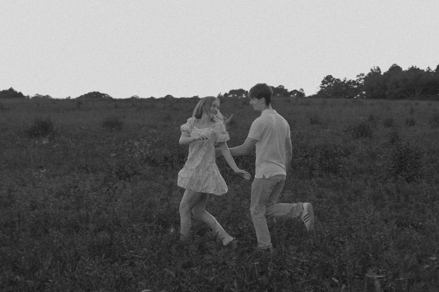 Black and white photo of a young couple holding hands and running through a lush green meadow with trees in background.