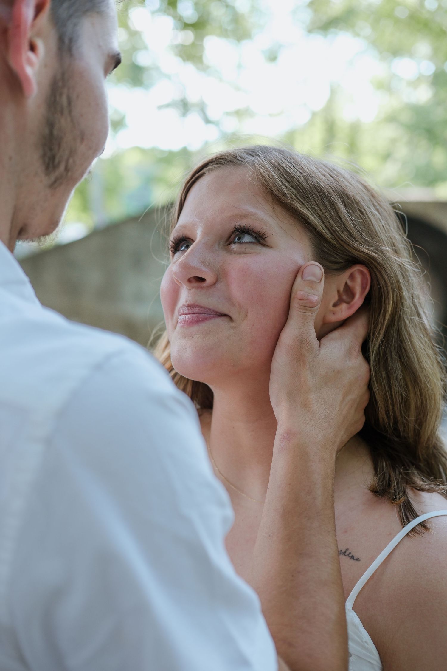 A man tenderly cups a smiling woman's face as she gazes up at him outdoors in a romantic moment.