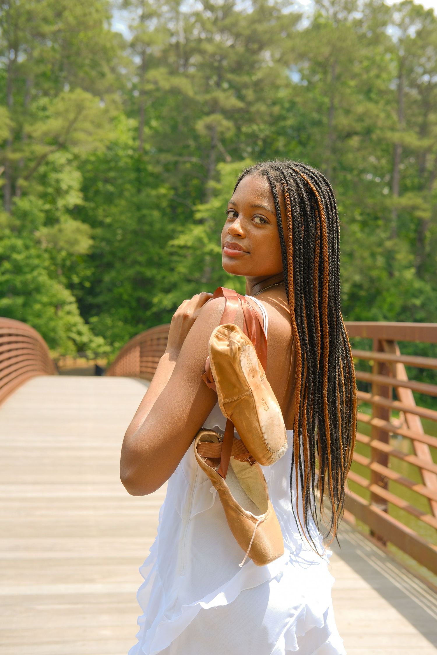 Young Black woman with long braids in white dress holds baguette bread on wooden bridge surrounded by green trees.