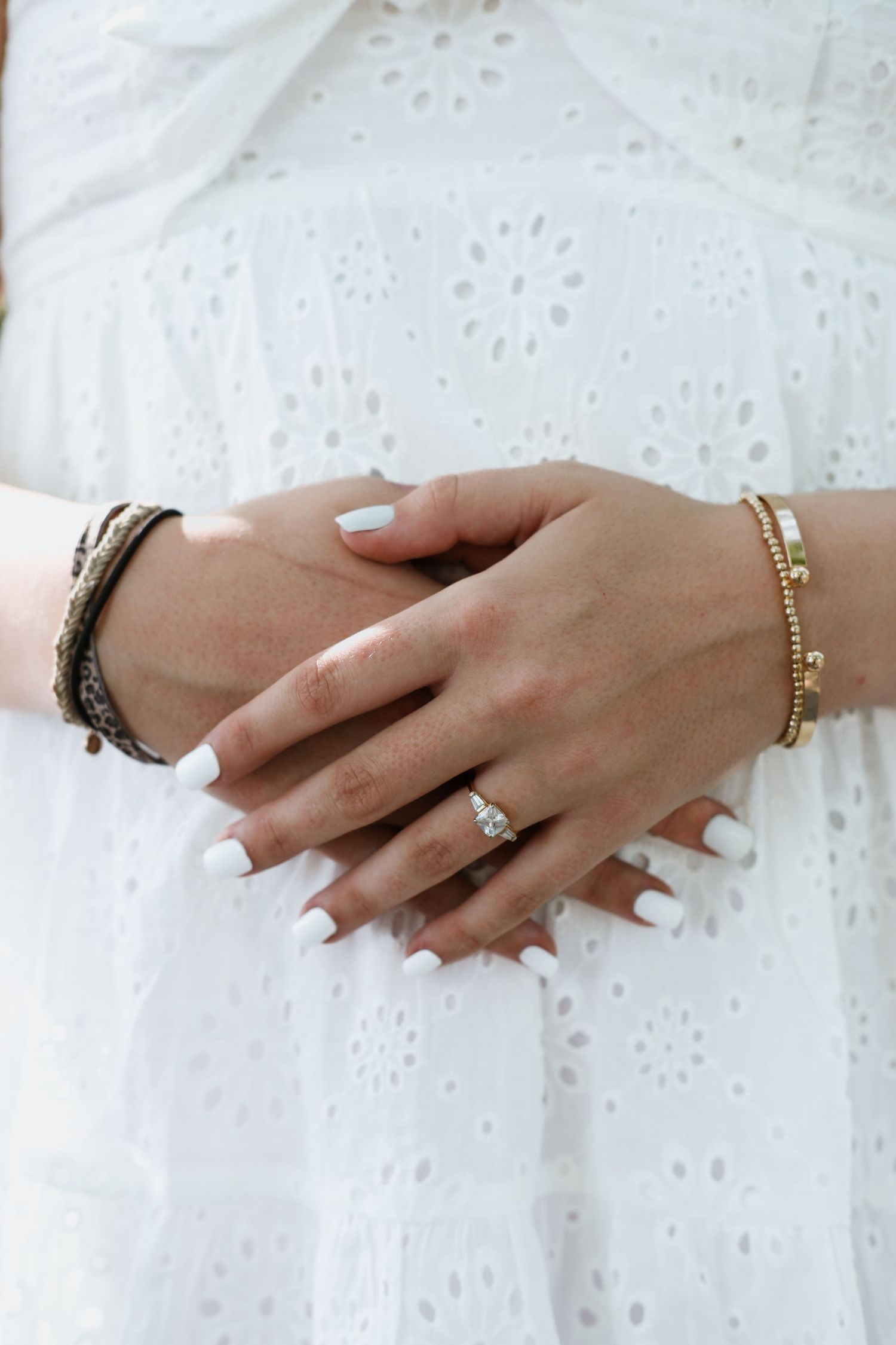 Close-up of clasped hands with white manicure, diamond ring, gold beaded bracelet, and woven bracelet on white eyelet dress.
