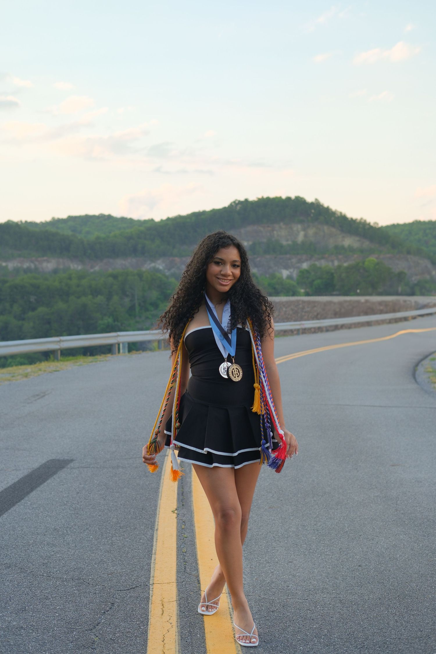 Young woman in black dress with medals stands on road with green hills and blue sky backdrop.