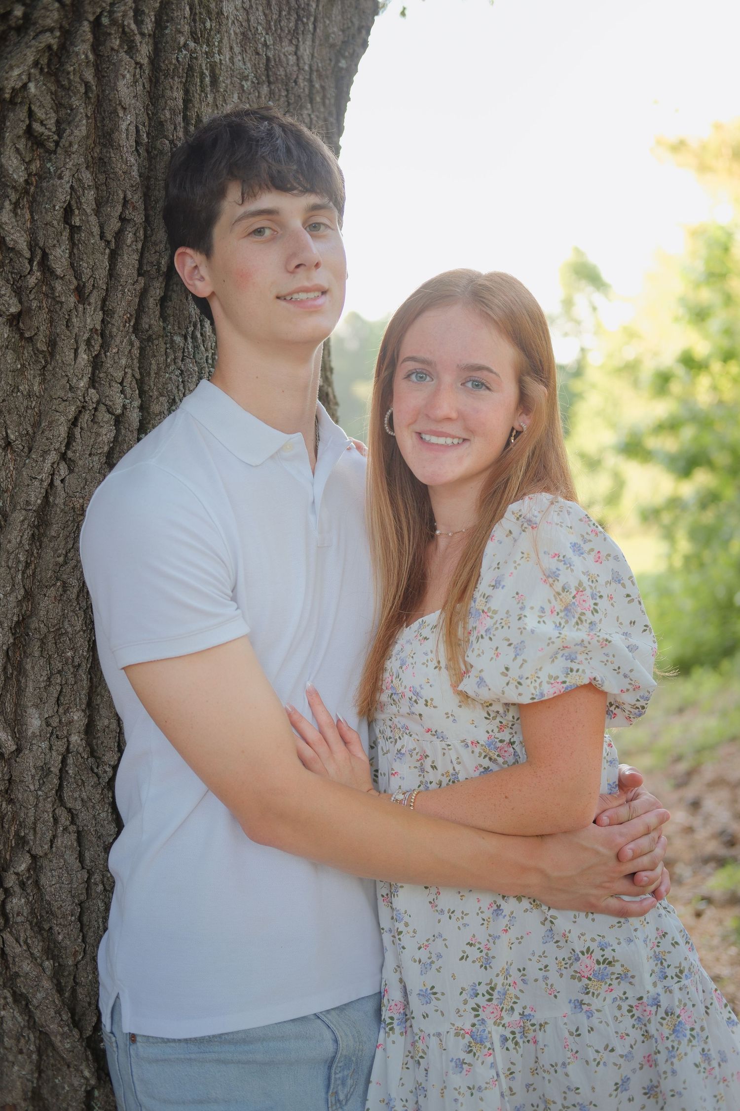 Young couple embracing by a large tree outdoors, smiling warmly in casual summer attire amid lush greenery.
