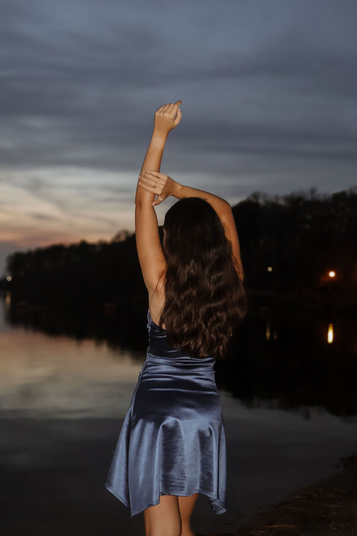 Woman in blue satin dress with arms raised facing serene lake at dusk.