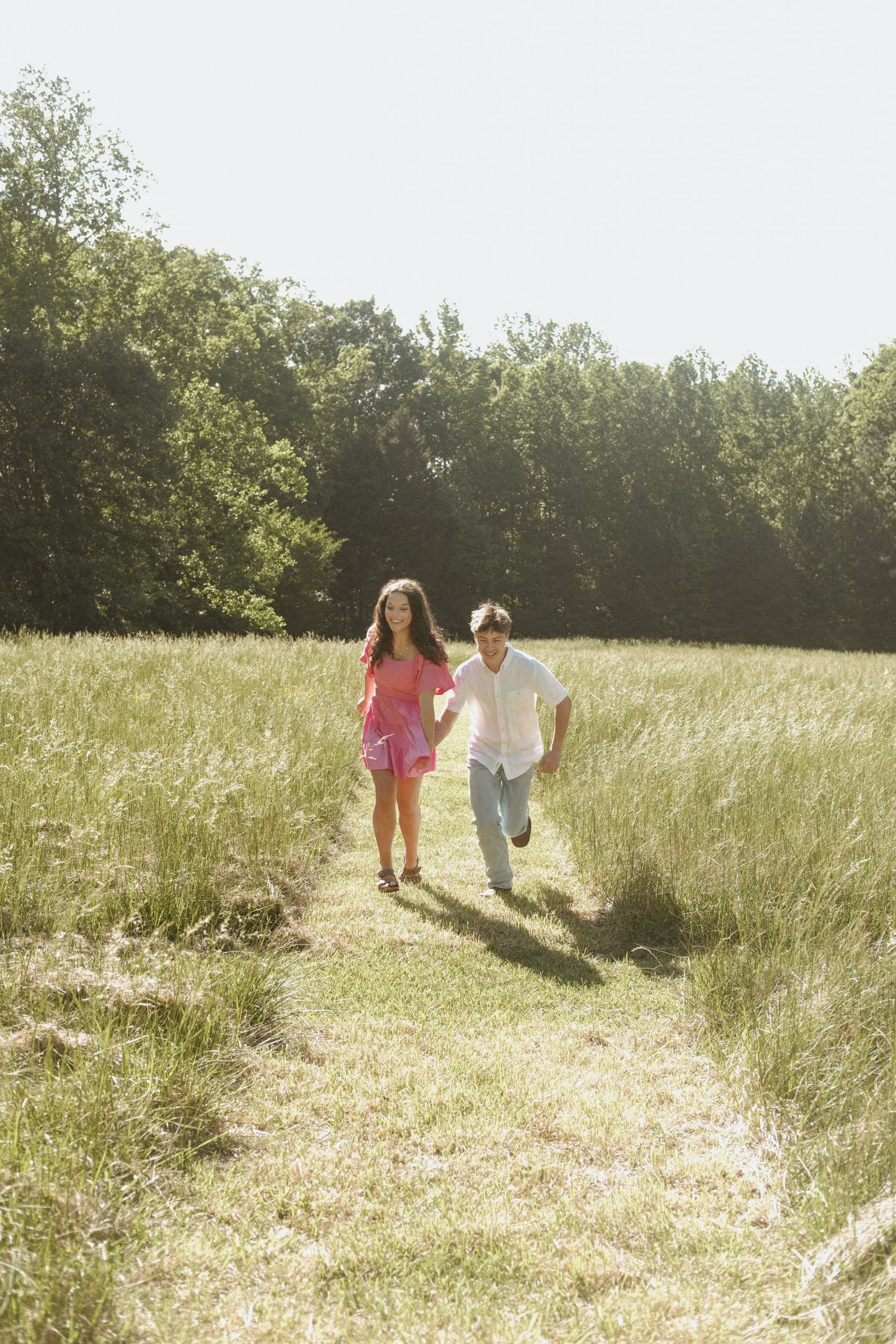 Young couple holding hands running through a sunny green meadow path with trees in background.