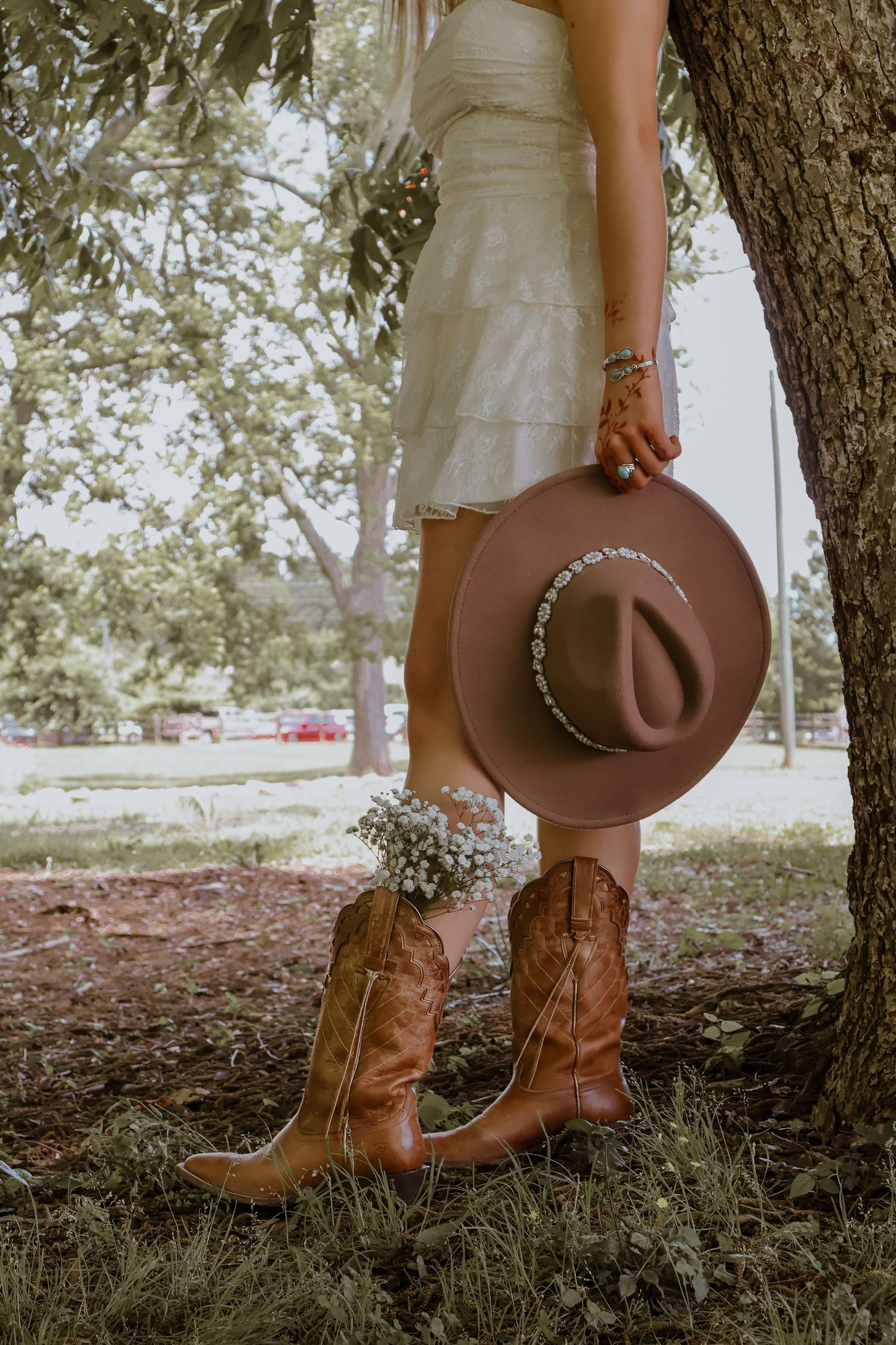 Woman in white dress holding brown cowboy hat with cowboy boots and wildflowers near a tree outdoors.