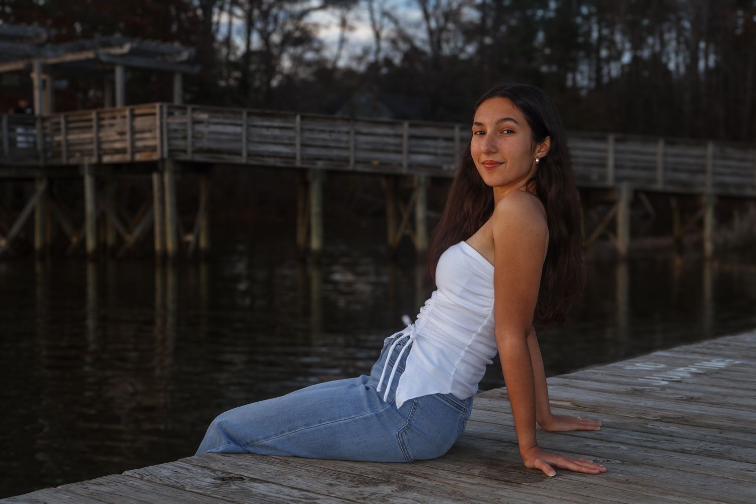 Young woman in white top and jeans sitting on a wooden dock by the water at dusk.