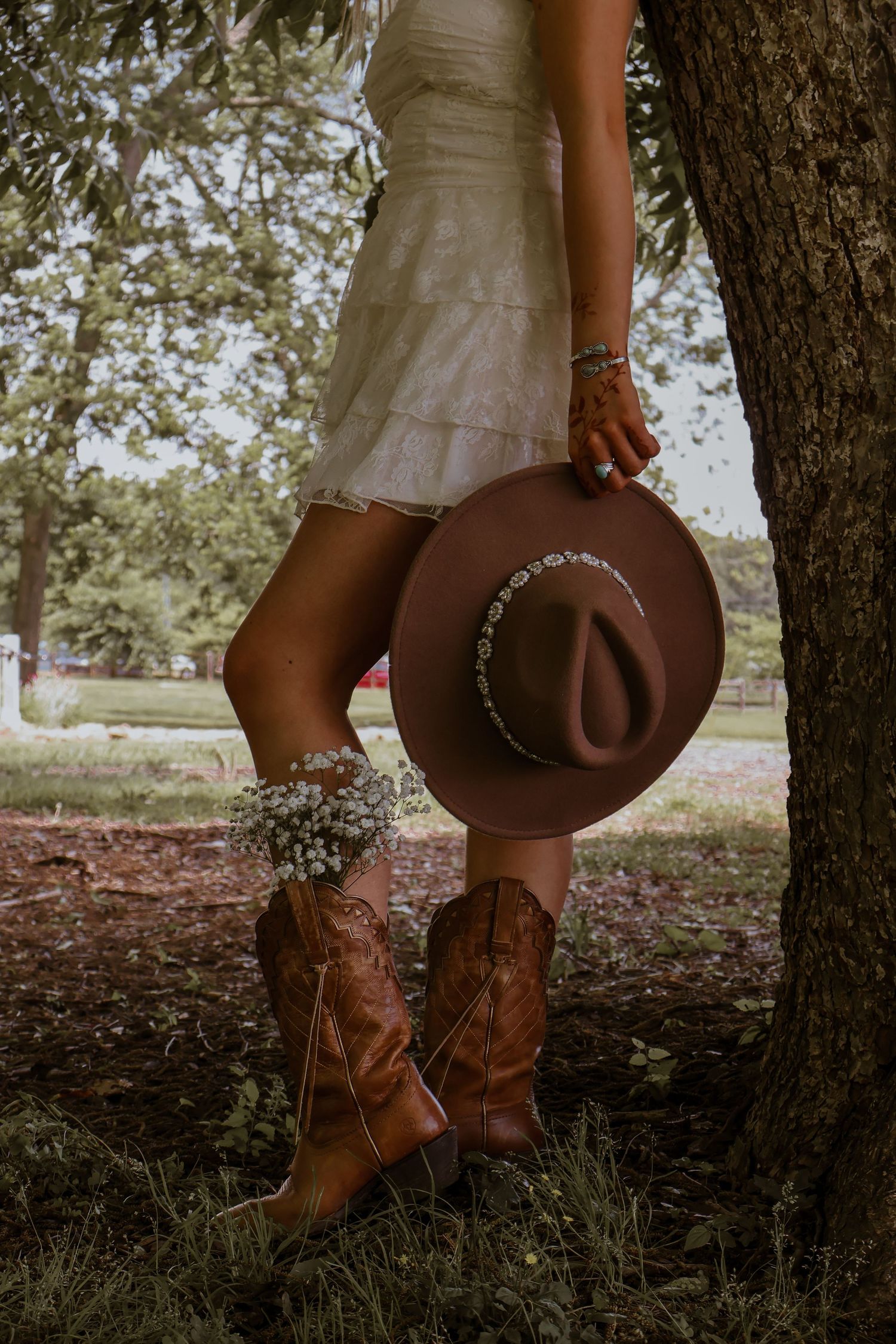 Close-up of woman in white dress holding brown cowboy hat wearing brown cowboy boots near tree.
