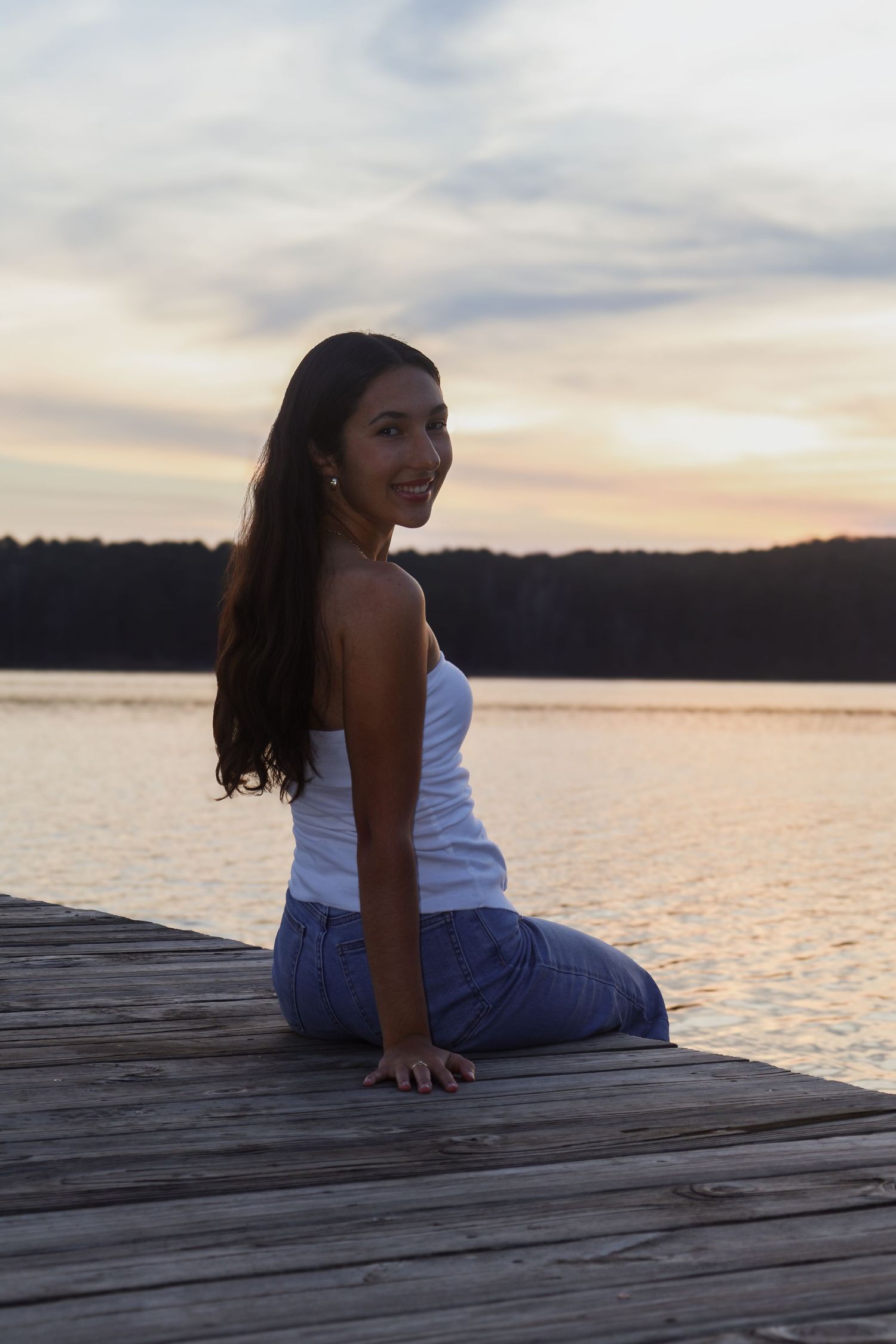 Smiling young woman in white top and jeans sitting on lakeside dock at sunset with colorful sky behind her.
