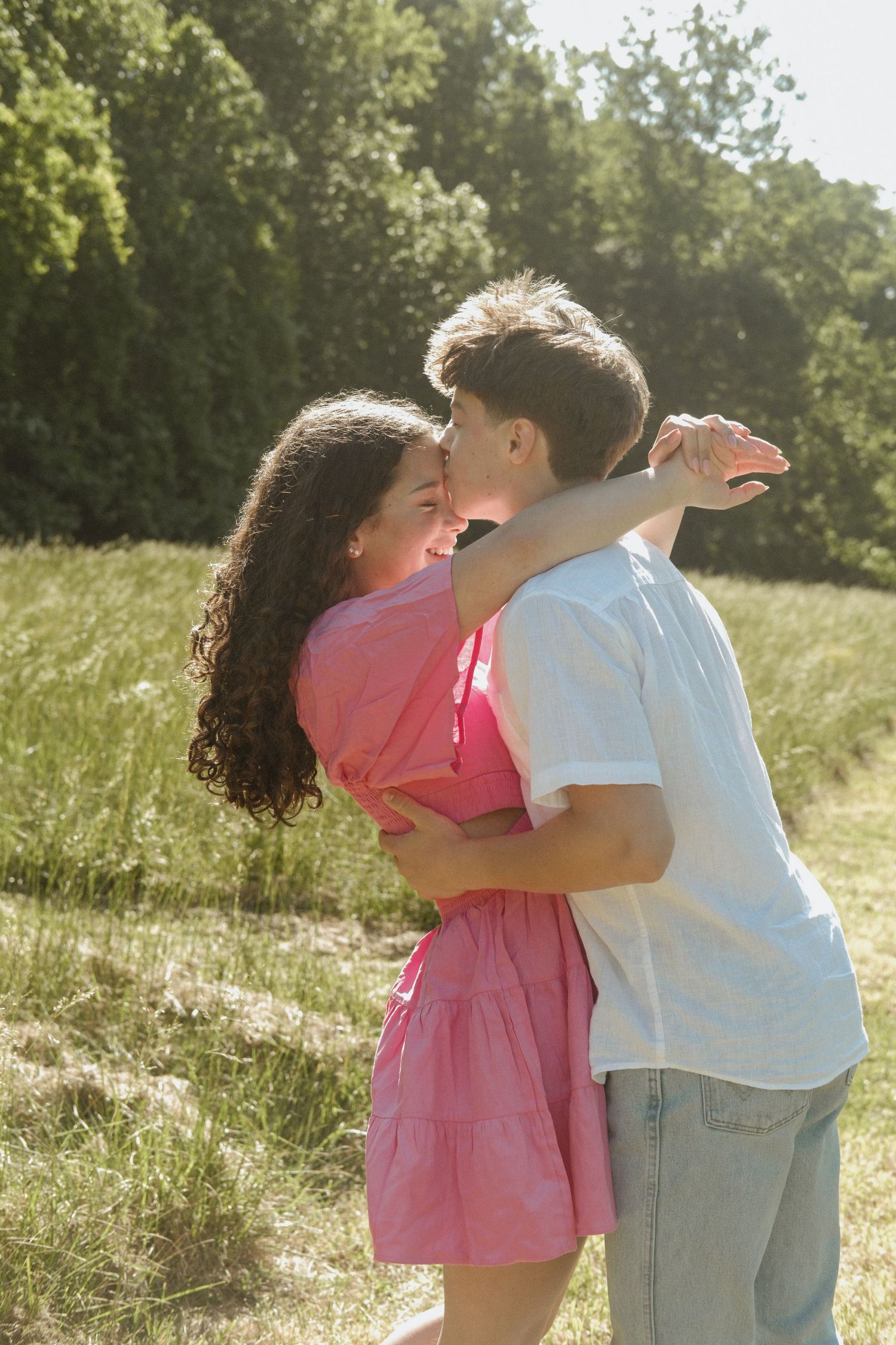 Young couple embracing and smiling in a sunny green meadow, woman in pink dress and man in white shirt.