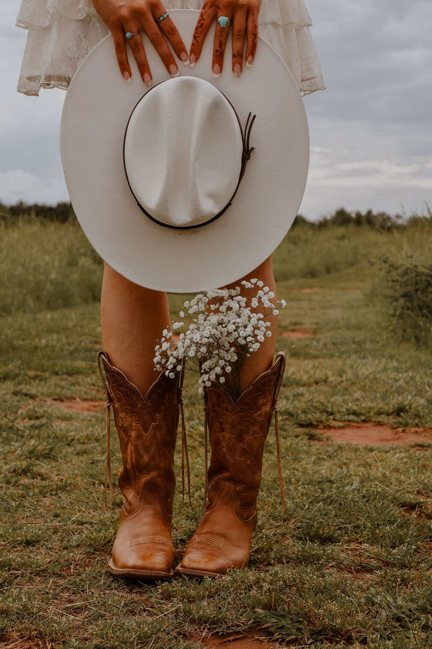 Woman in white dress holding white wide-brim hat with cowboy boots and baby's breath flowers outdoors.