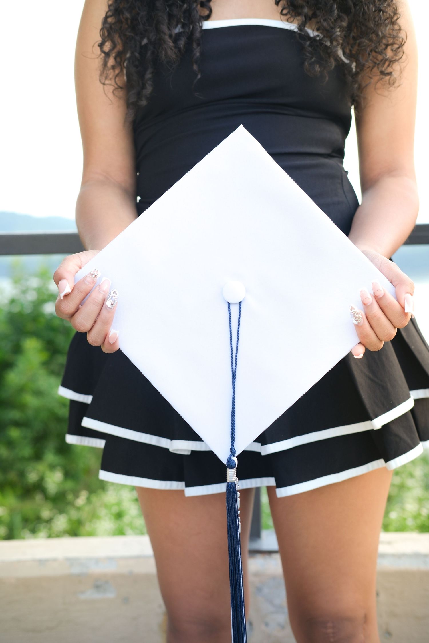 Young woman in black dress holding white graduation cap with blue tassel outdoors.